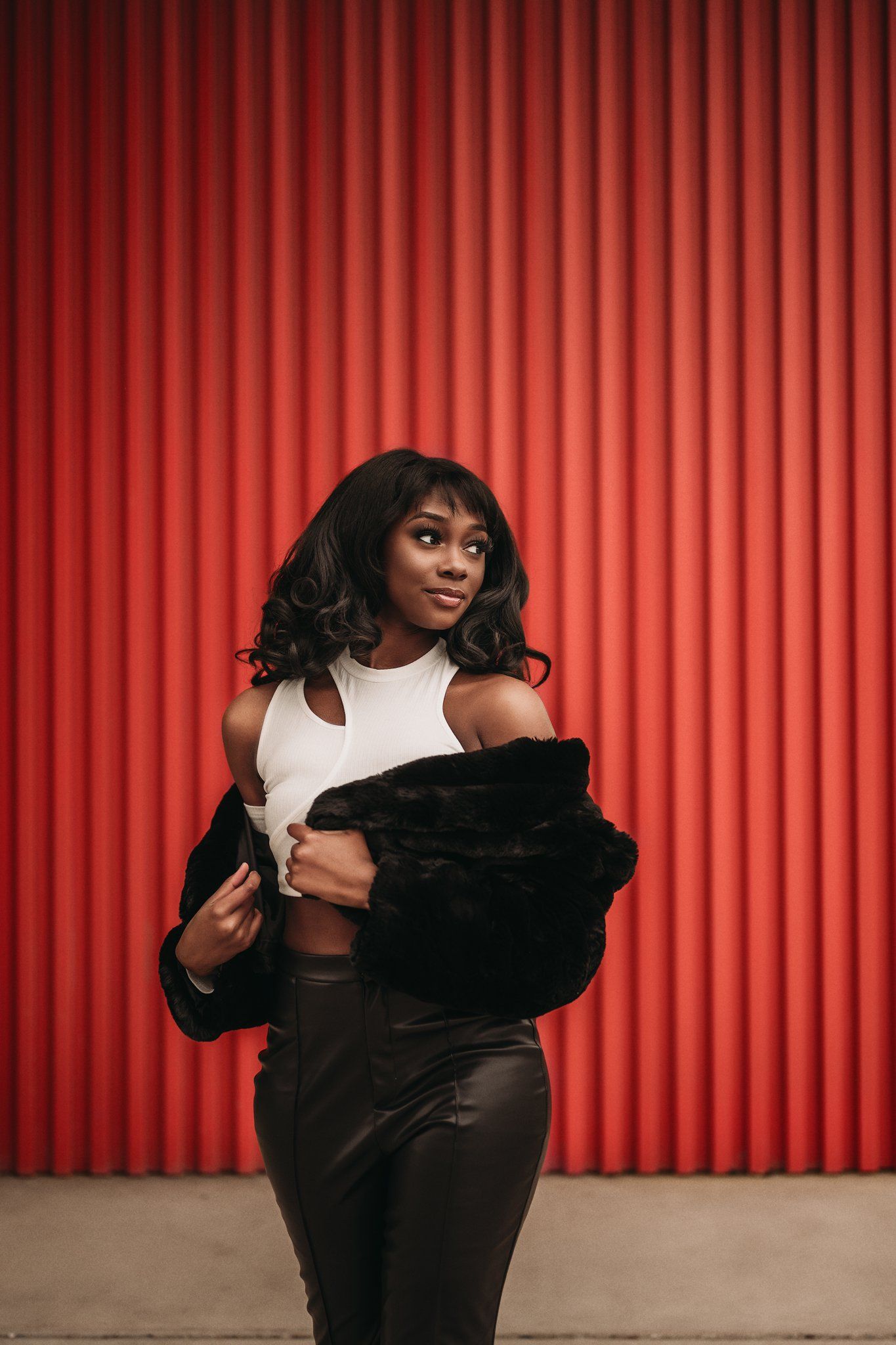 A model strikes a playful pose wearing a white top and black fur coat against red stripes.