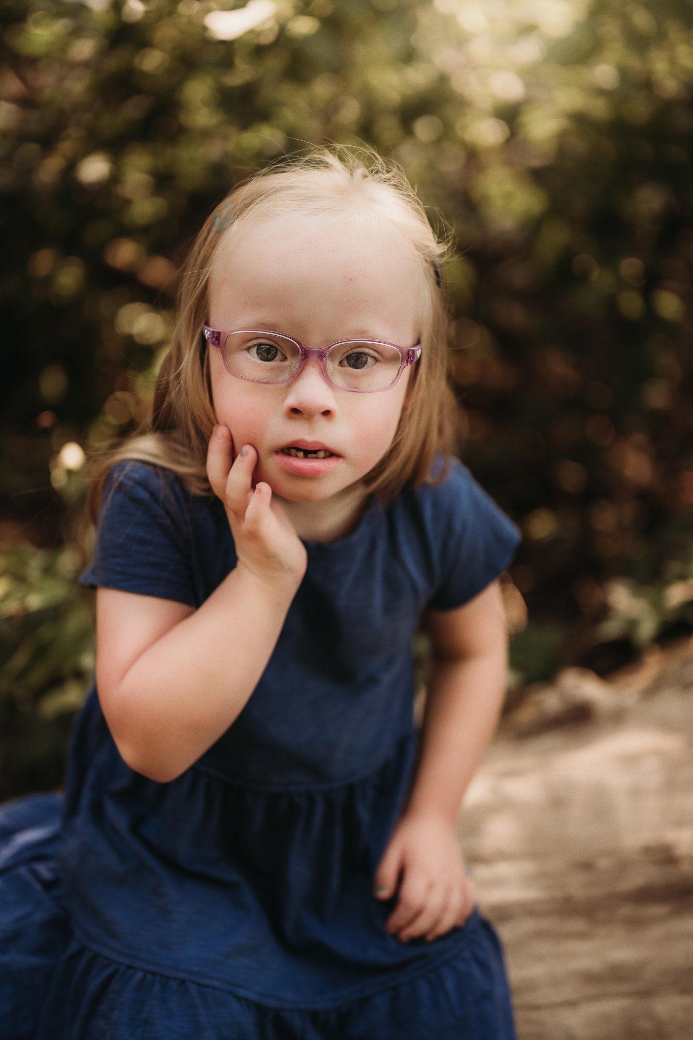 A thoughtful child in a navy dress and glasses sits on a wooden log outdoors.