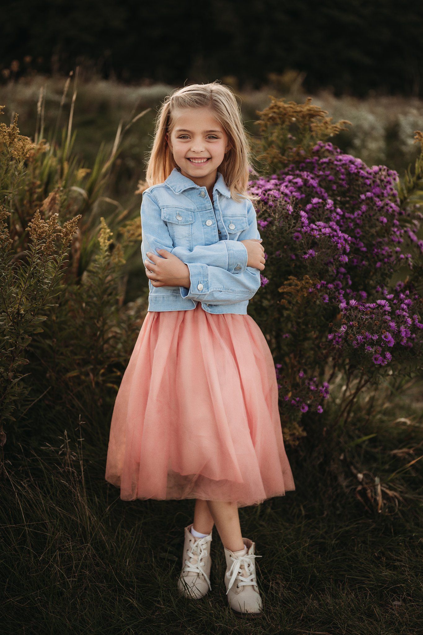 Young person in denim jacket and pink tulle skirt poses confidently among purple flowers.