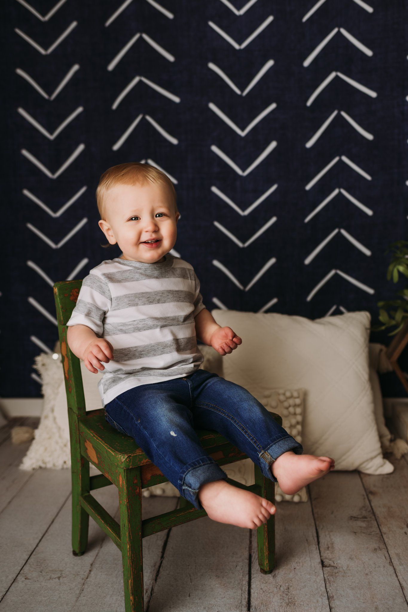 A toddler wearing a gray striped shirt sits happily on a green wooden chair.