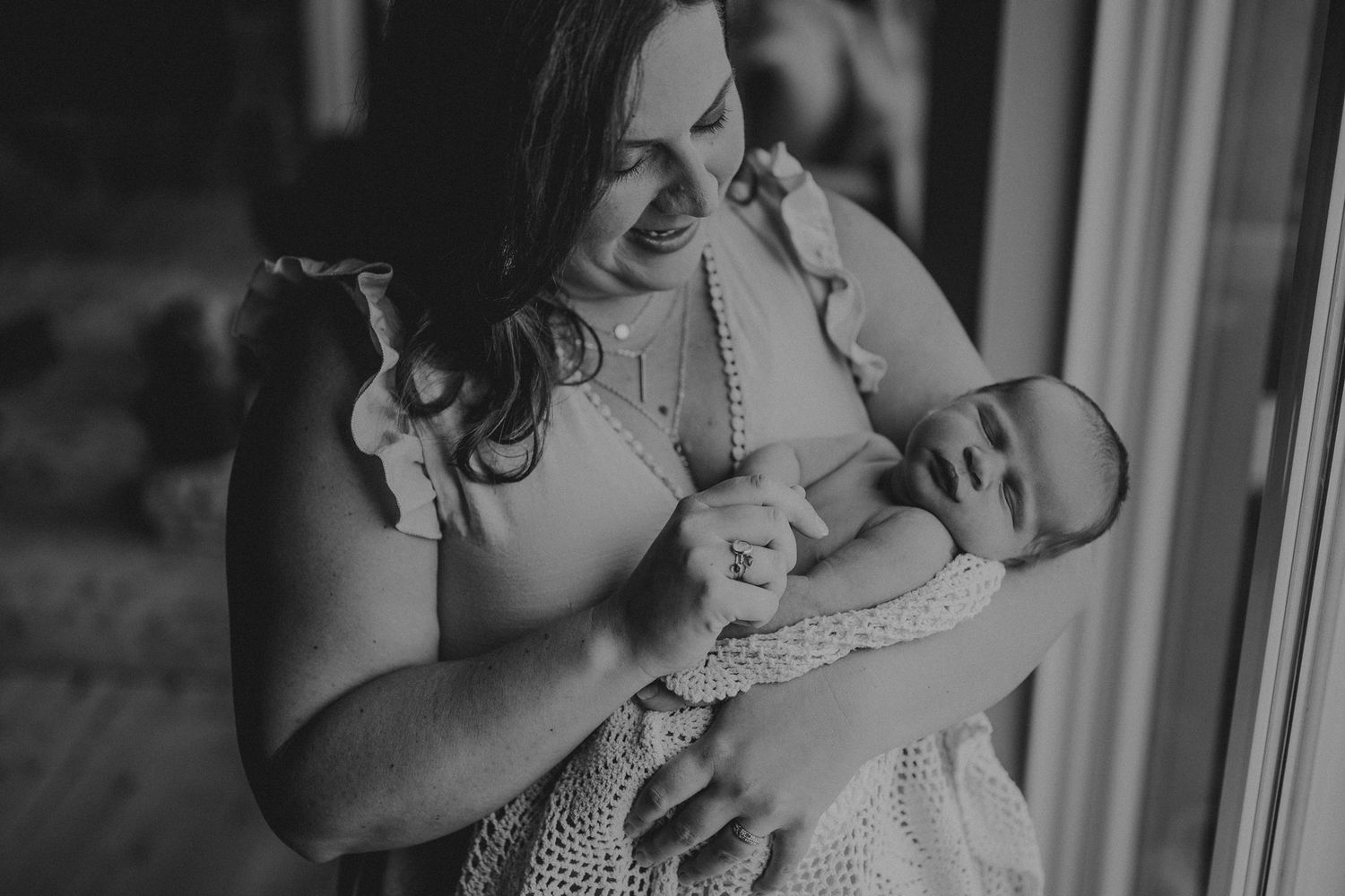 Black and white portrait of maternal bonding as baby rests against lace dress near window.