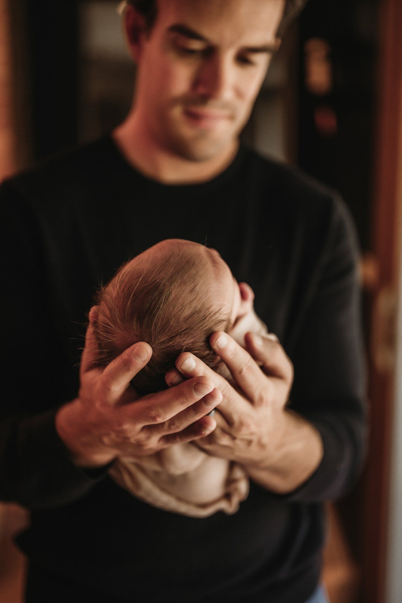 Person wearing black gently holds sleeping newborn baby in atmospheric lighting.