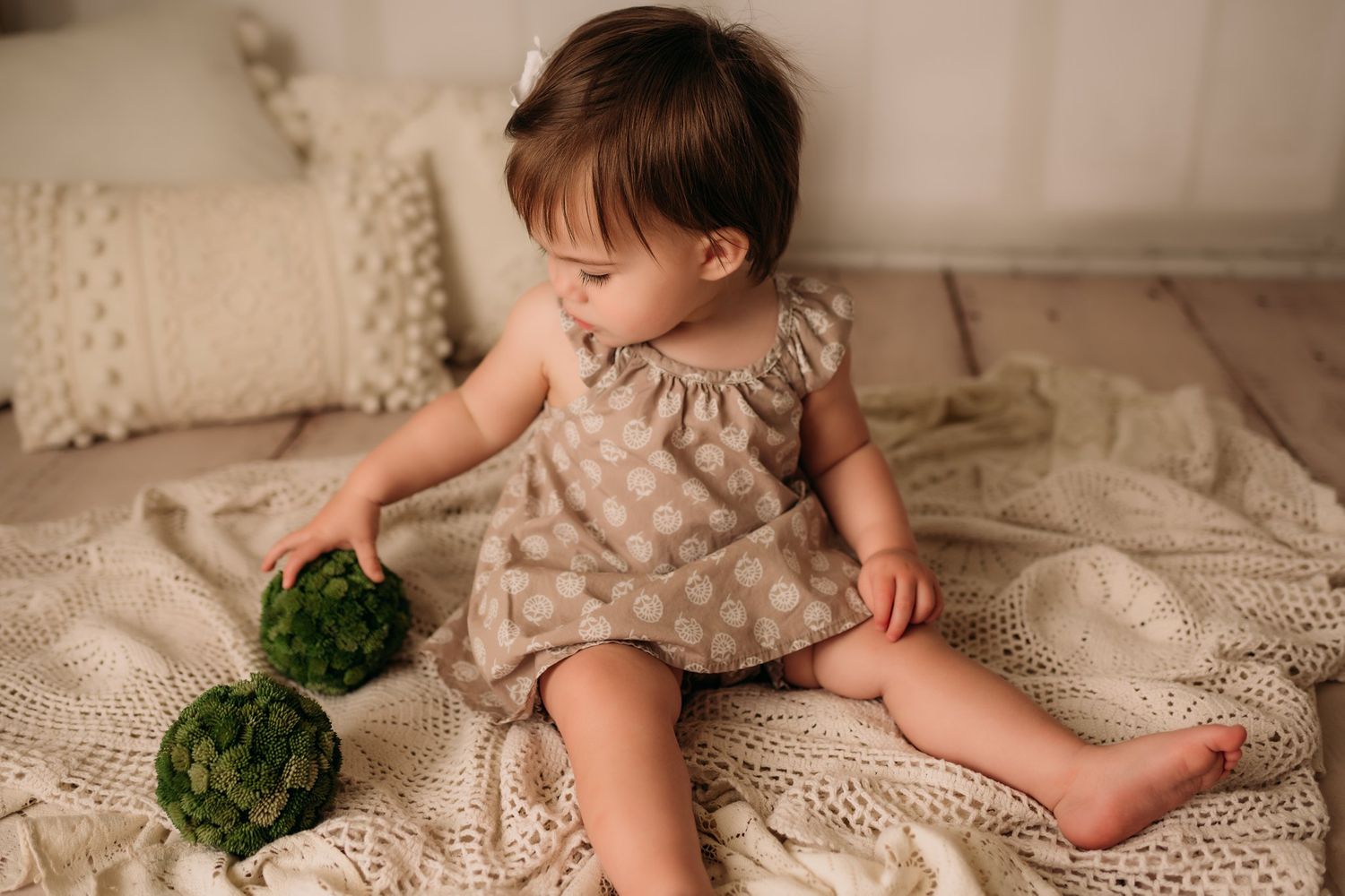 Baby in beige dress playing with green moss balls on cream textured blankets.