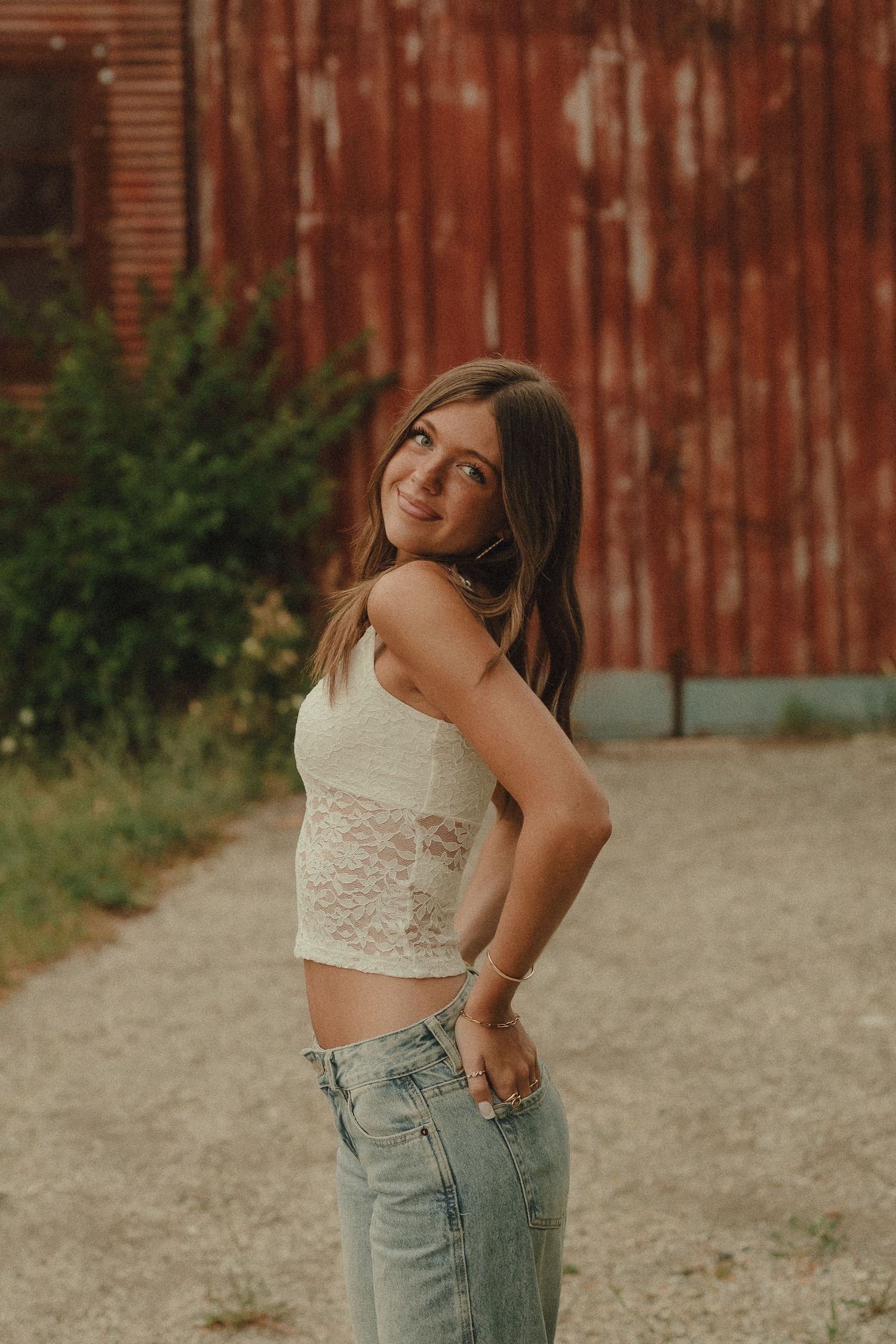 Teenage girl in white lace crop top and light wash jeans smiling beside a rustic red barn.