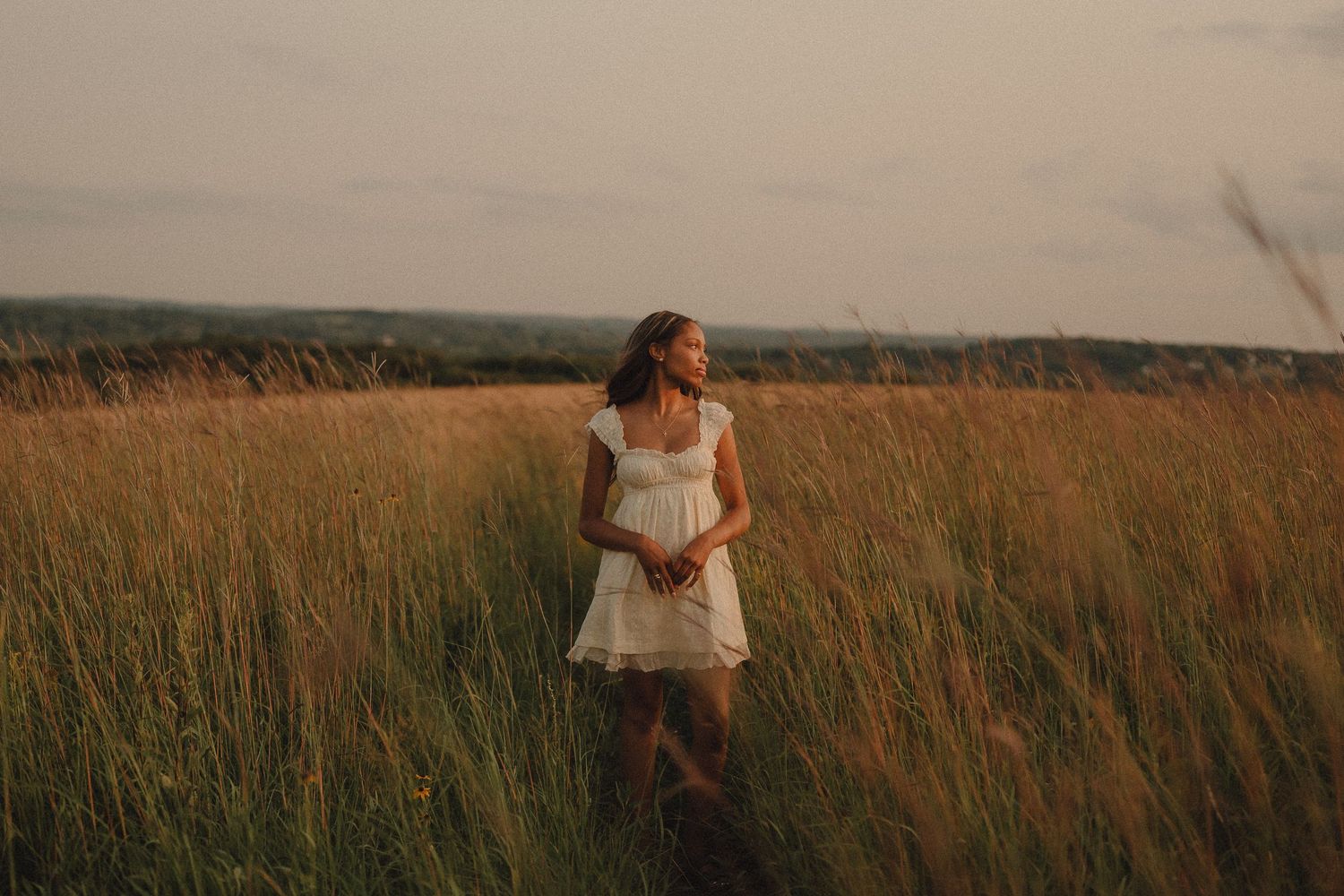 A young woman in a white dress stands peacefully in a tall golden grass field at dusk.