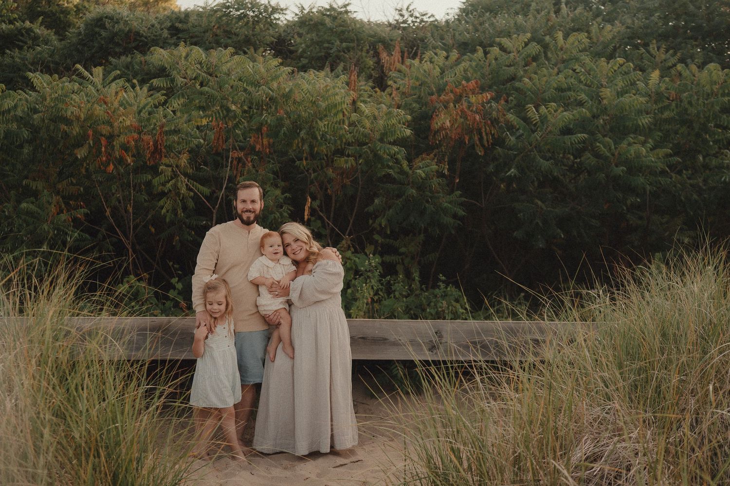 A happy family of four poses together on a sandy path surrounded by tall beach grass.