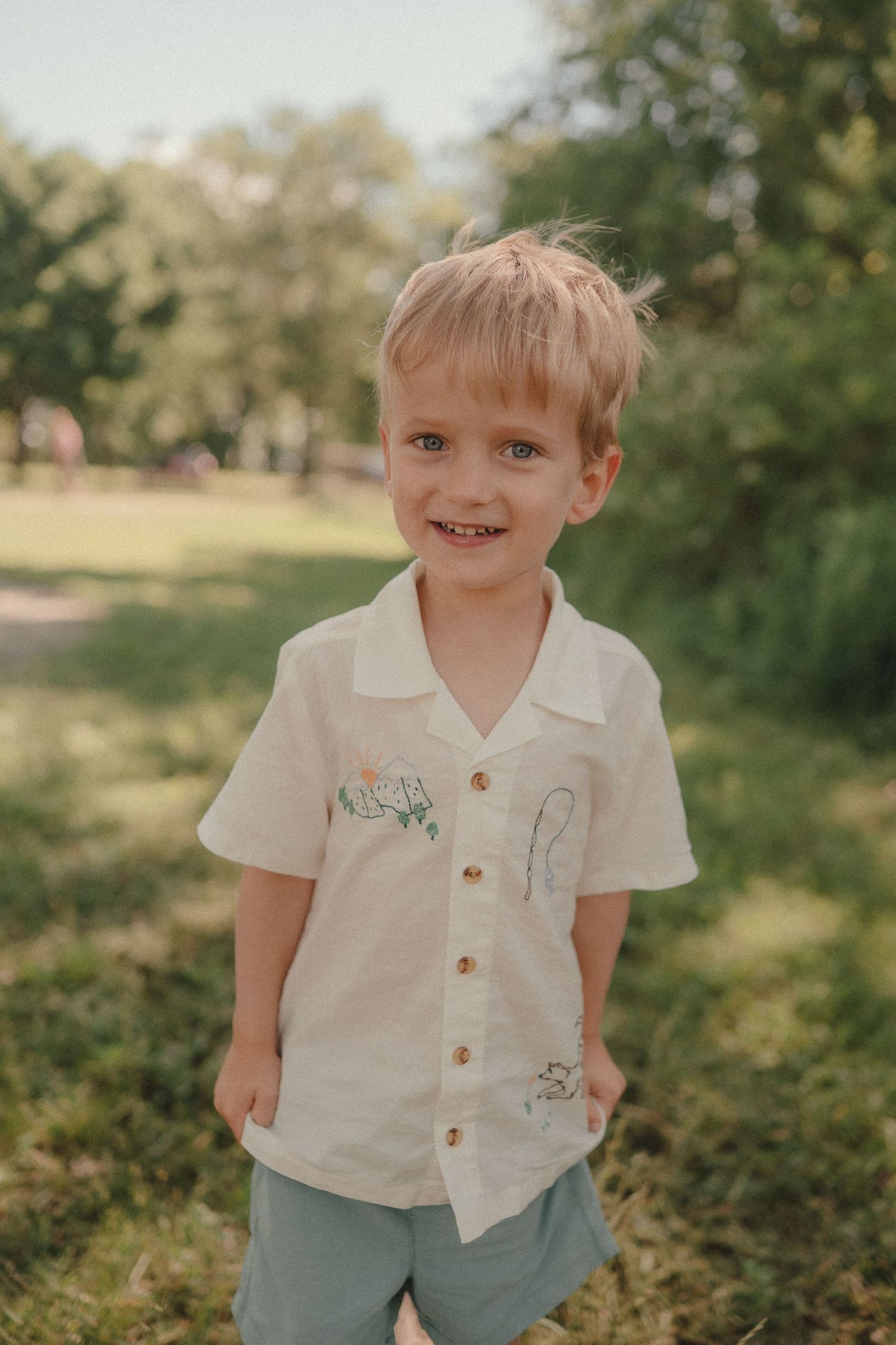 Smiling young blonde boy in a white embroidered button-up shirt standing outdoors in a green park setting.