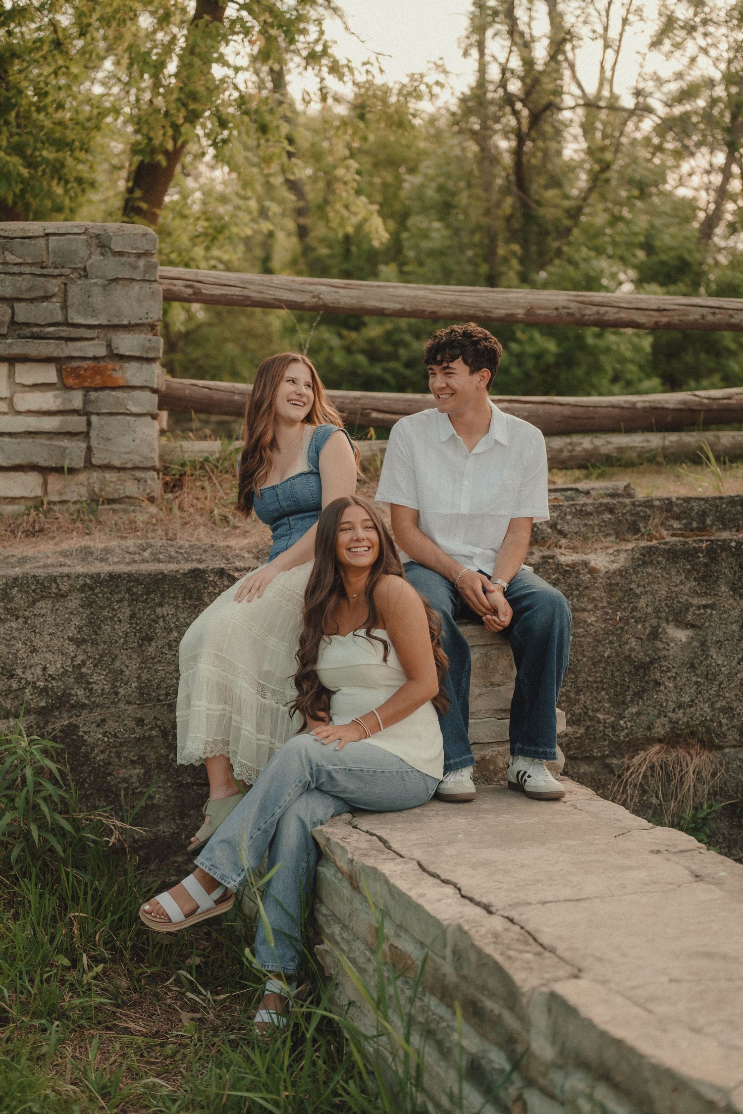 Three smiling teens sit on a stone wall surrounded by lush green trees outdoors.
