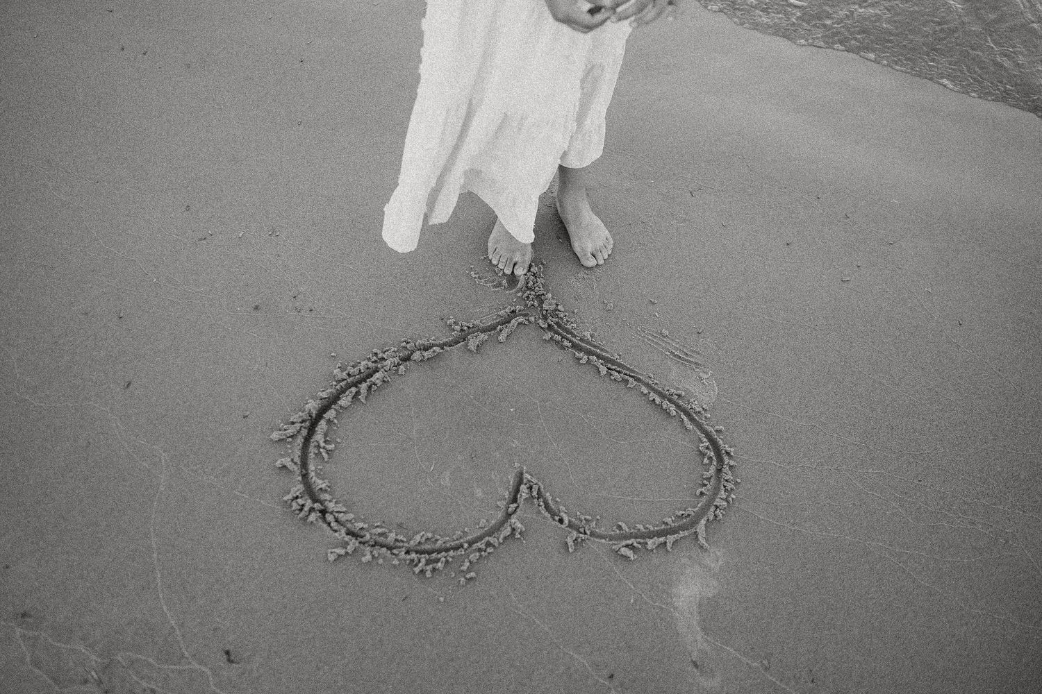 Heart drawn in wet sand on beach with woman in white dress standing nearby, black and white photo.