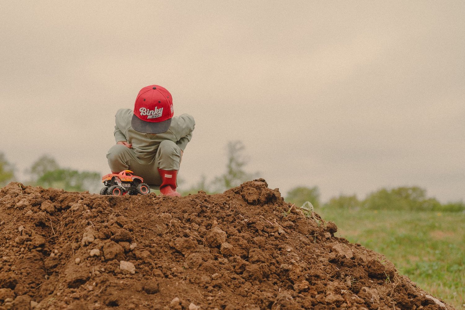 Young child in red cap playing on a large dirt mound outdoors on a cloudy day.