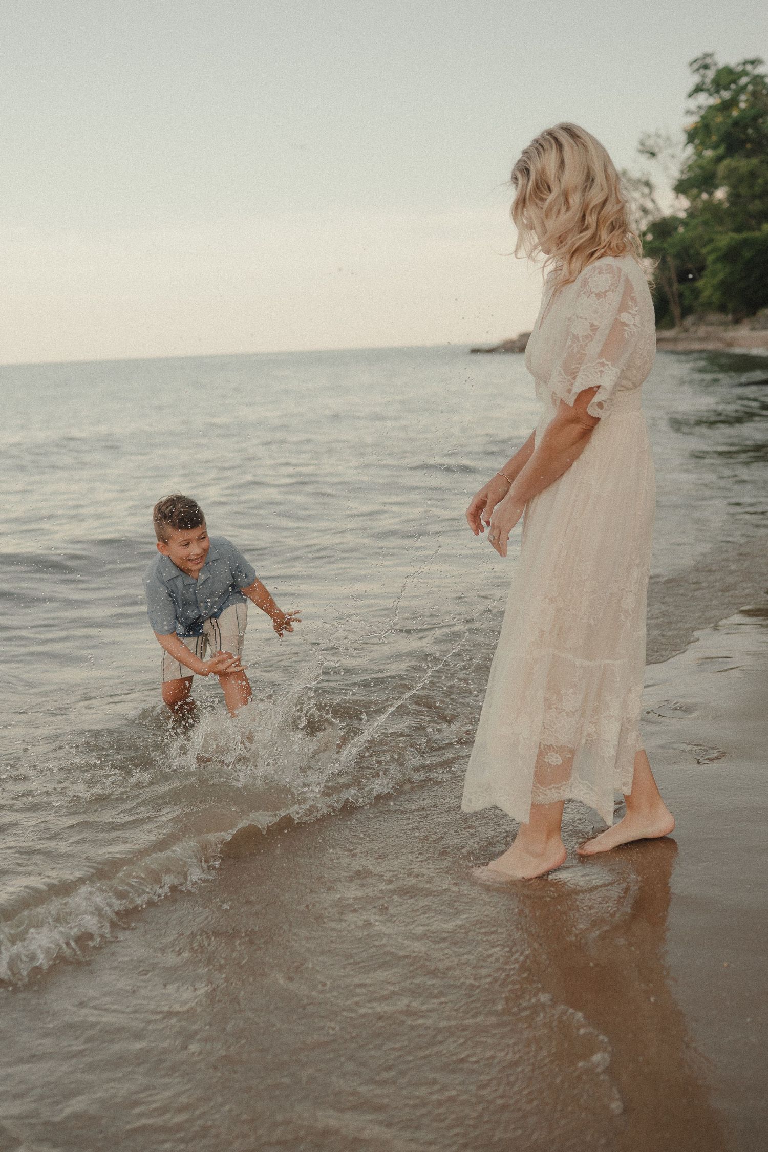 A woman in a white lace dress and a young boy play in ocean waves on a sandy beach at sunset.