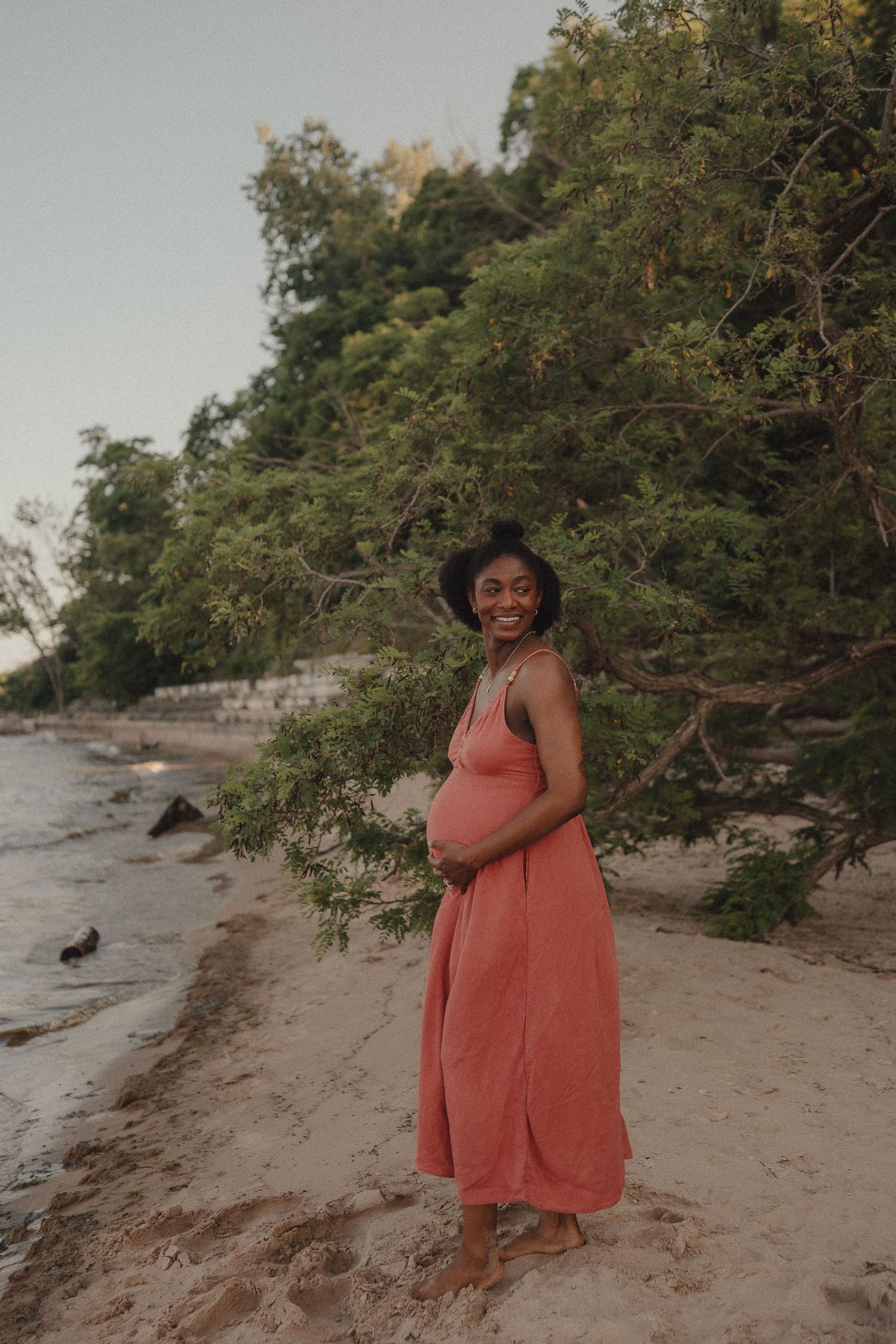Smiling pregnant Black woman in coral maxi dress standing barefoot on sandy beach near green trees at dusk.