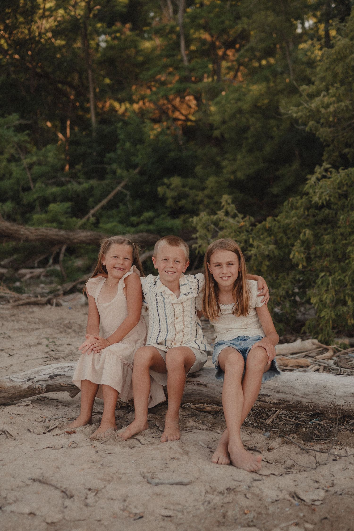 Three barefoot siblings sit together on driftwood on a sandy beach, smiling warmly surrounded by lush green trees.