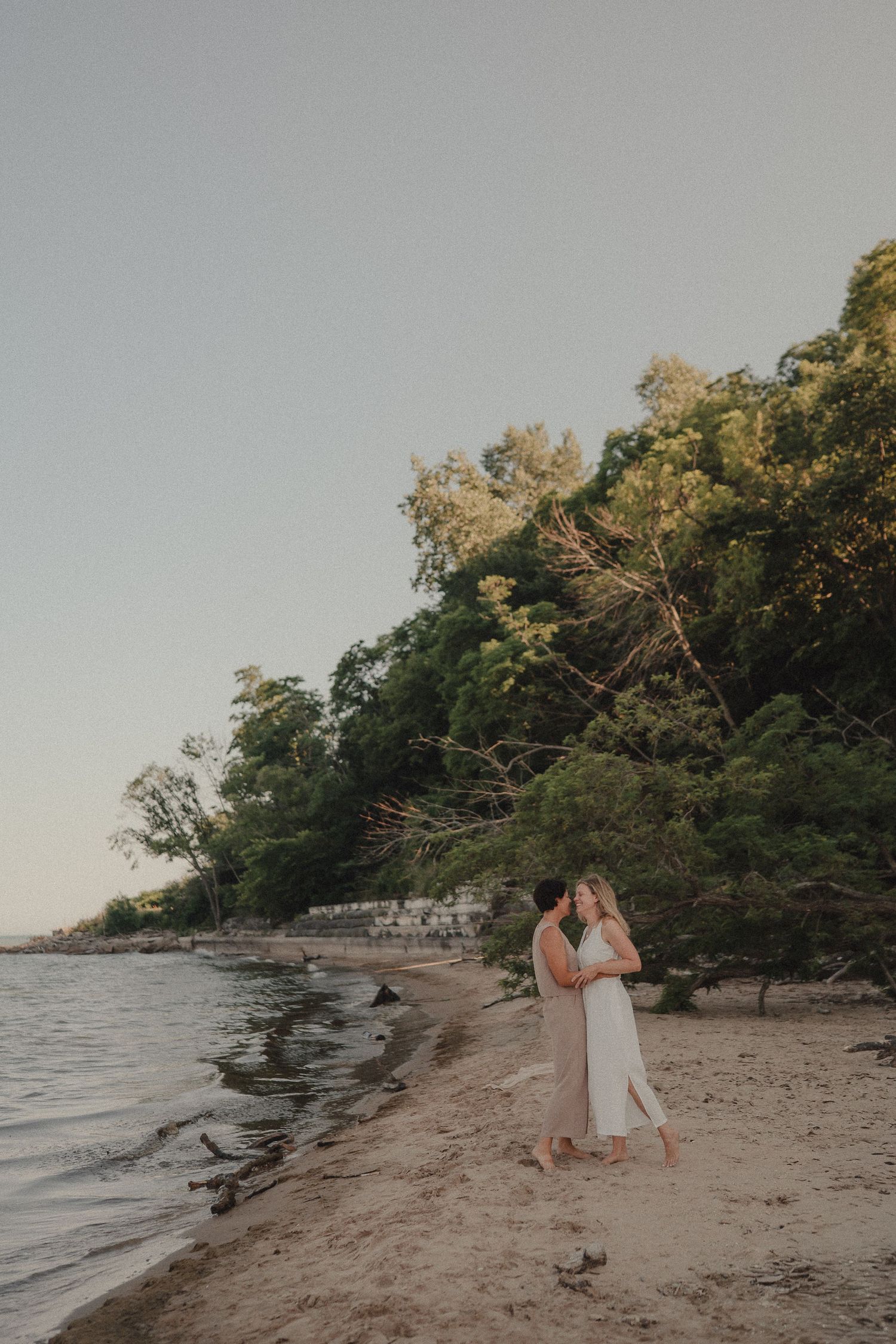Couple sharing a romantic kiss on a sandy beach at dusk with lush trees behind.