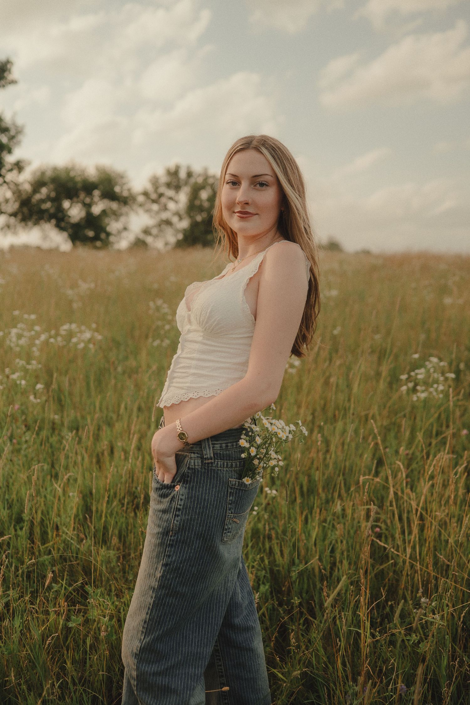Smiling young woman in white crop top and jeans holding wildflowers in a grassy meadow.
