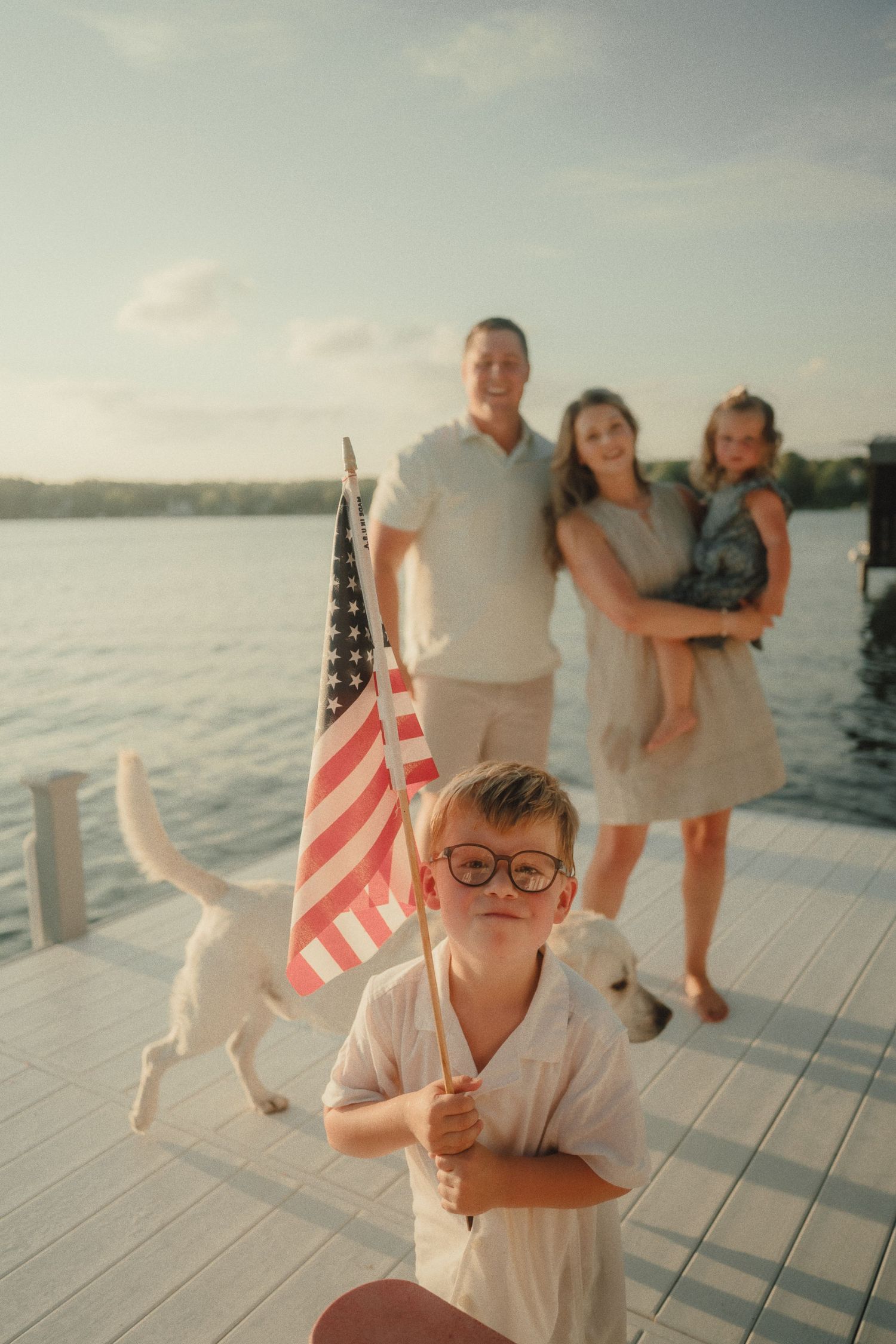A boy in glasses holds an American flag on a dock while his family and dog pose by the lake behind him.
