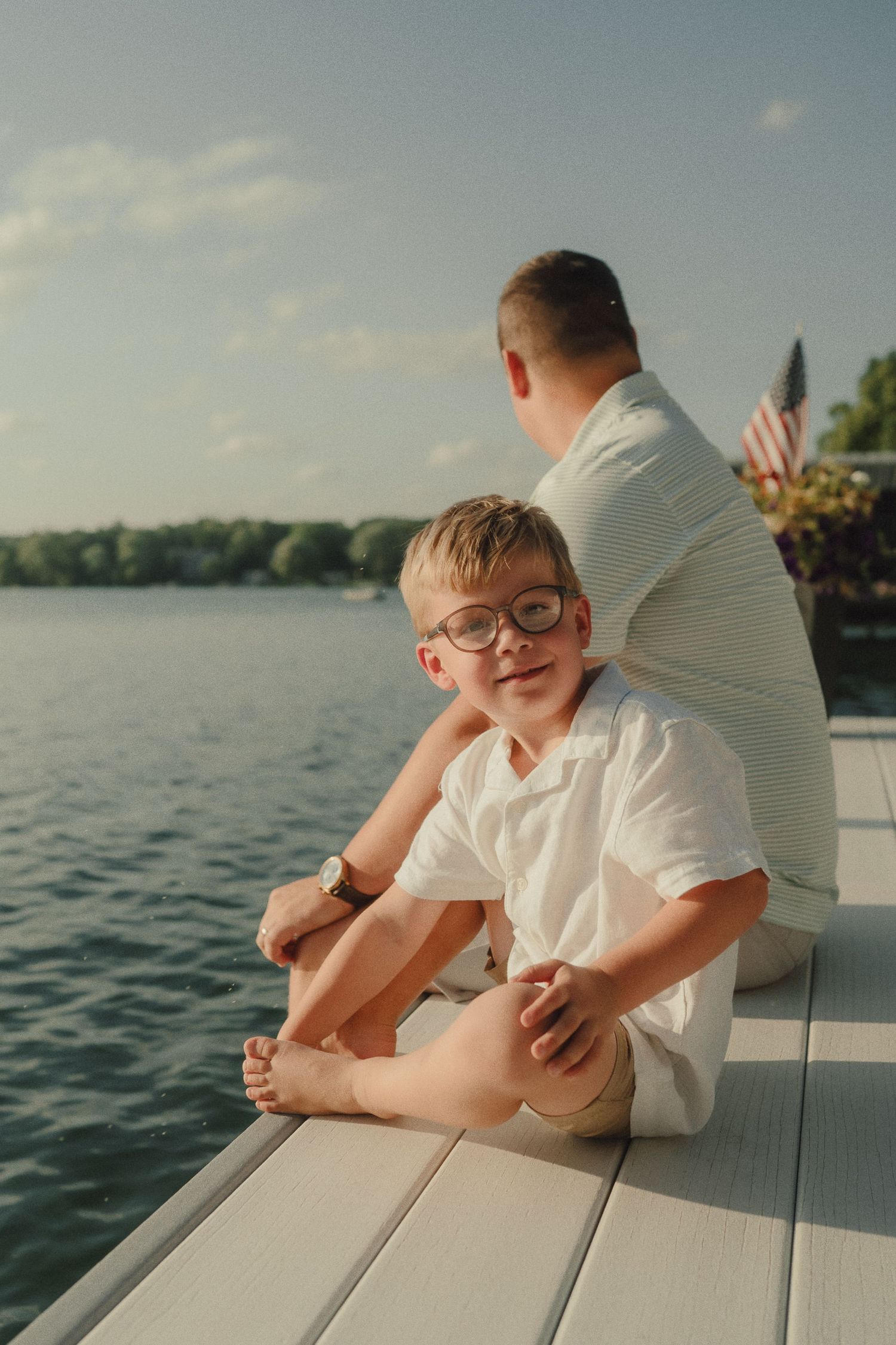 A young boy in glasses and white shirt sits on a dock by a lake, smiling at camera, with an adult beside him.