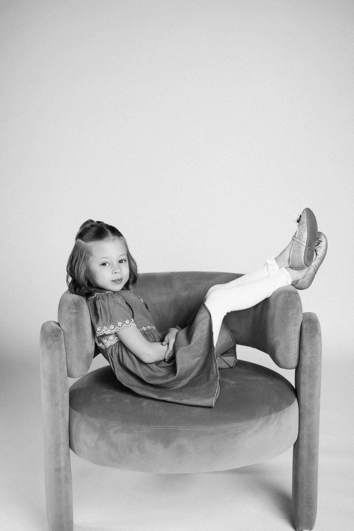 A child lounges casually in a plush velvet armchair wearing a dress and white shoes in a black and white portrait.