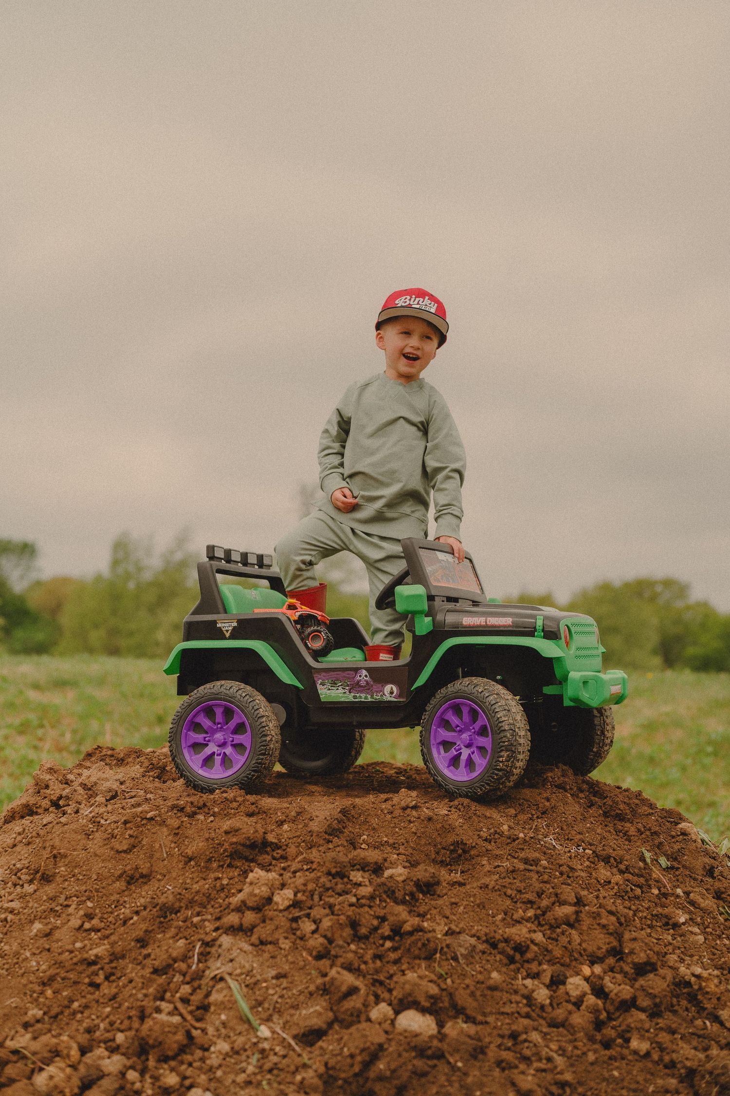 Smiling boy in red cap standing atop green toy jeep on dirt mound outdoors.