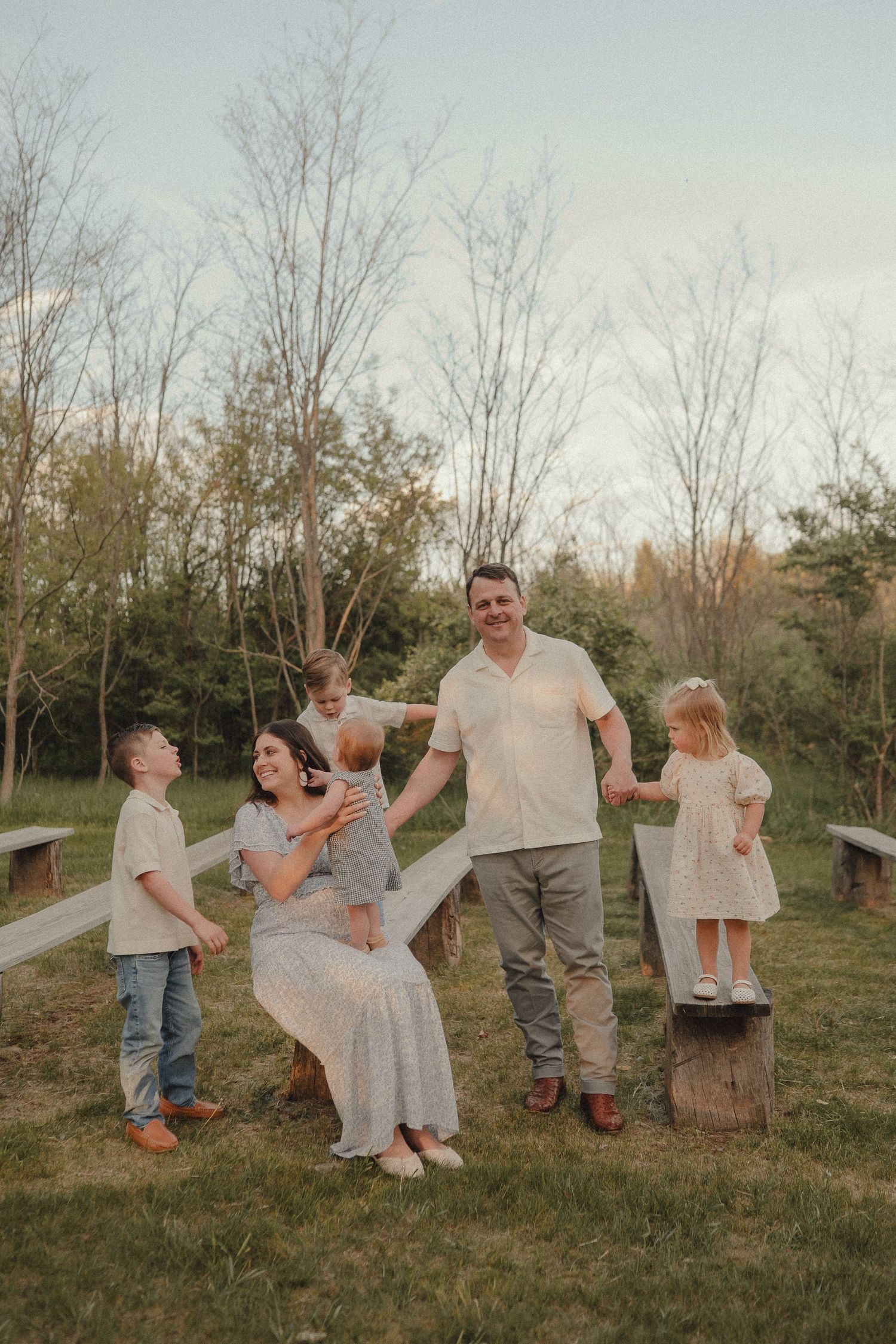 A joyful family of five plays together on a grassy lawn with wooden benches at golden hour.