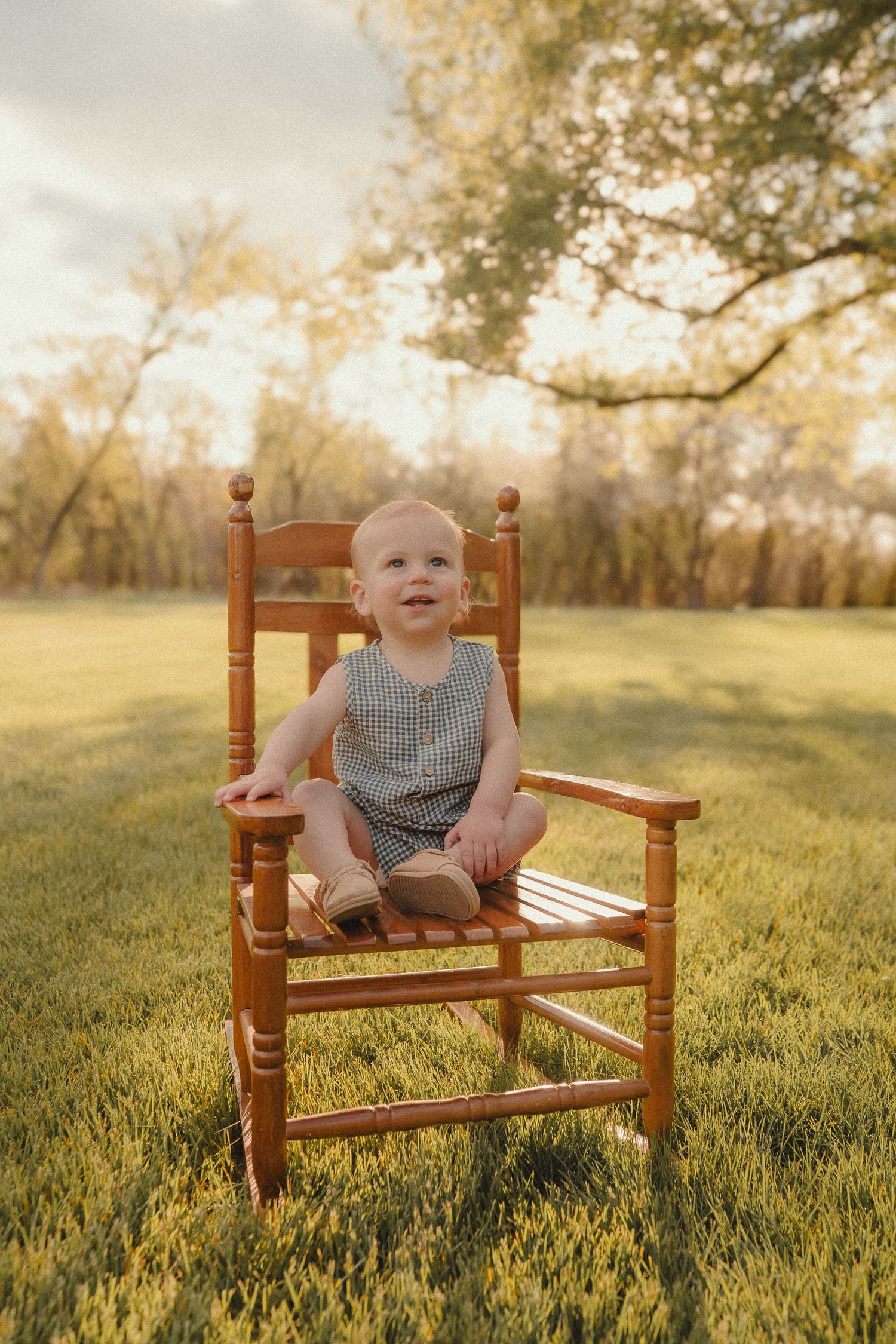 Smiling baby in a gingham romper sitting in a wooden chair on green grass at golden hour.