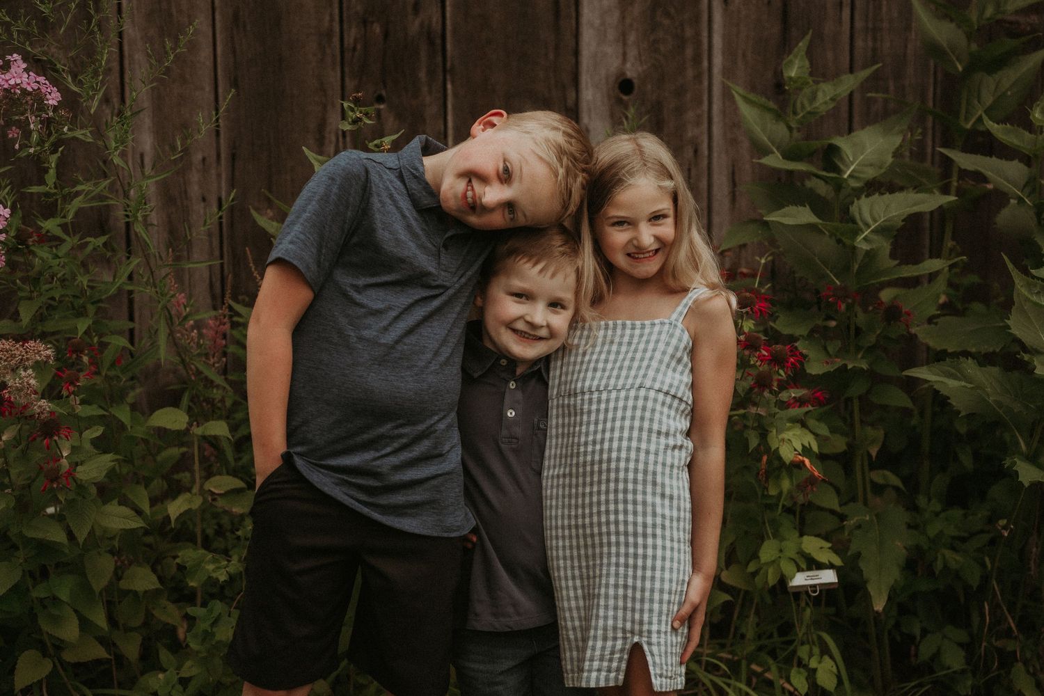 Three children pose together against rustic barn wall with flowering garden plants.