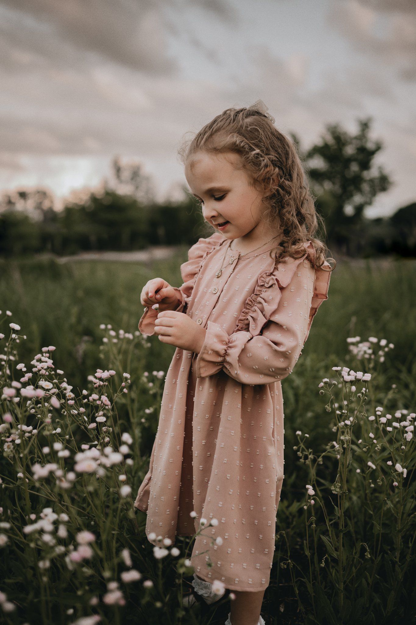 Child in vintage-style dress examines flowers in a meadow at dusk.