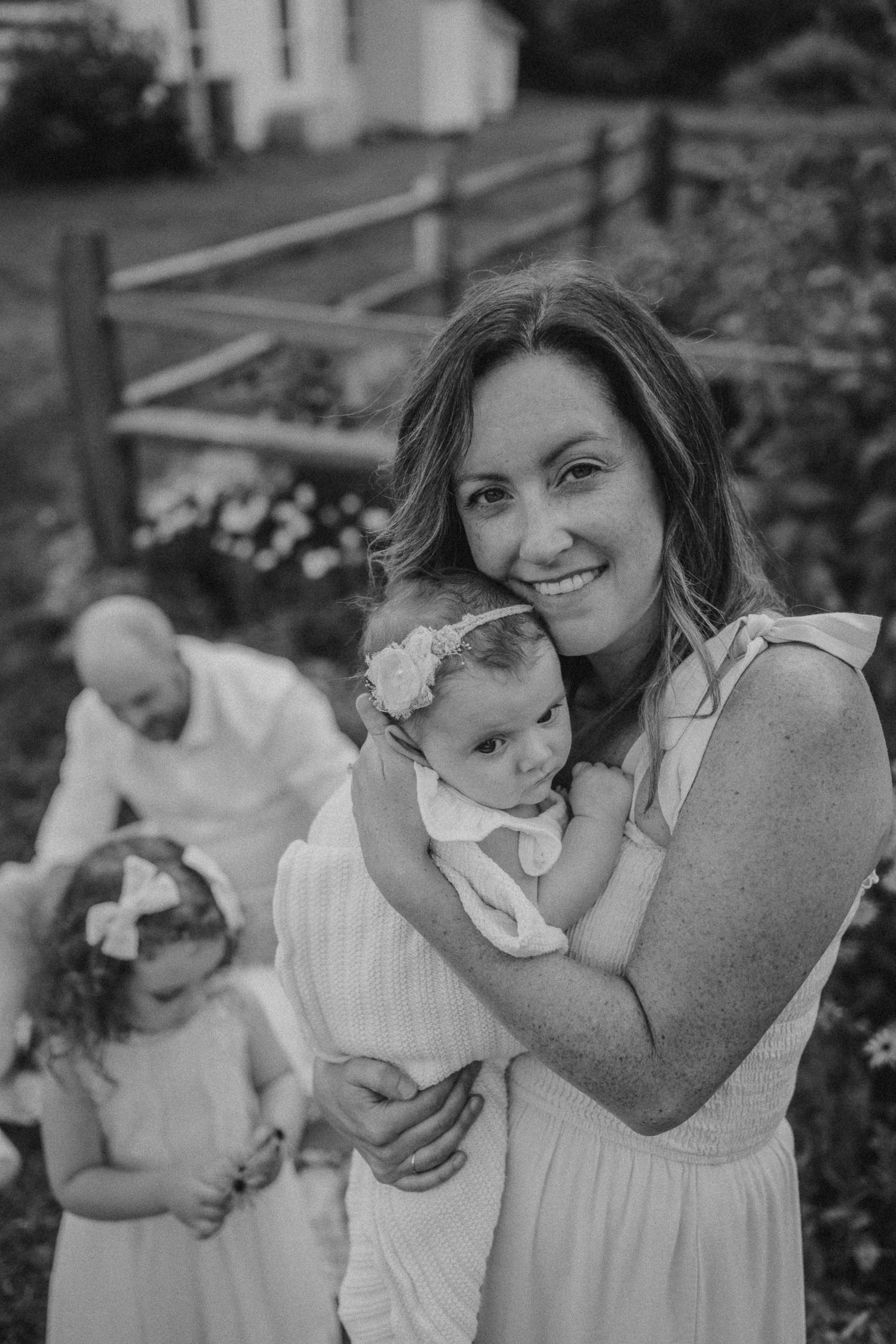 A black and white portrait of a family moment in a garden setting with a rustic fence.