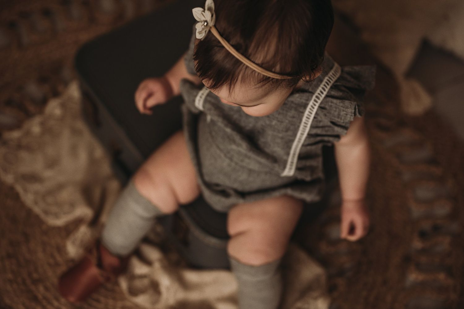 Baby in grey romper and knee socks plays on patterned rug in moody lighting.
