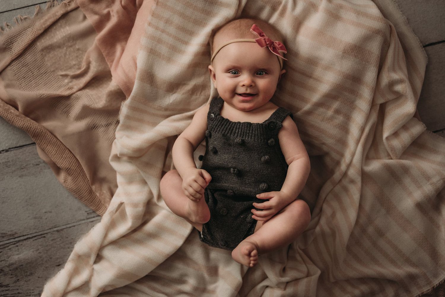 Baby wearing dark romper smiling while lying on blush colored fabric.