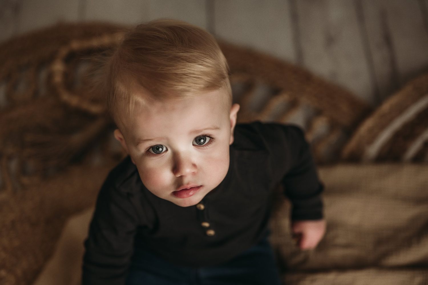 Moody portrait of a toddler in black henley sitting against vintage wooden furniture.