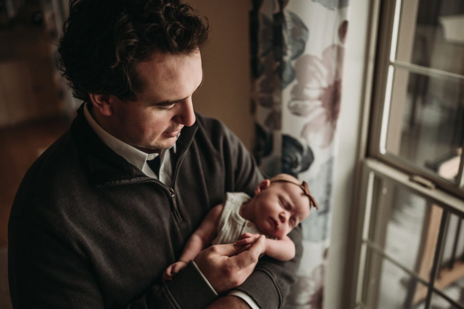 Parent standing by window in dark sweater holding and studying their newborn baby.