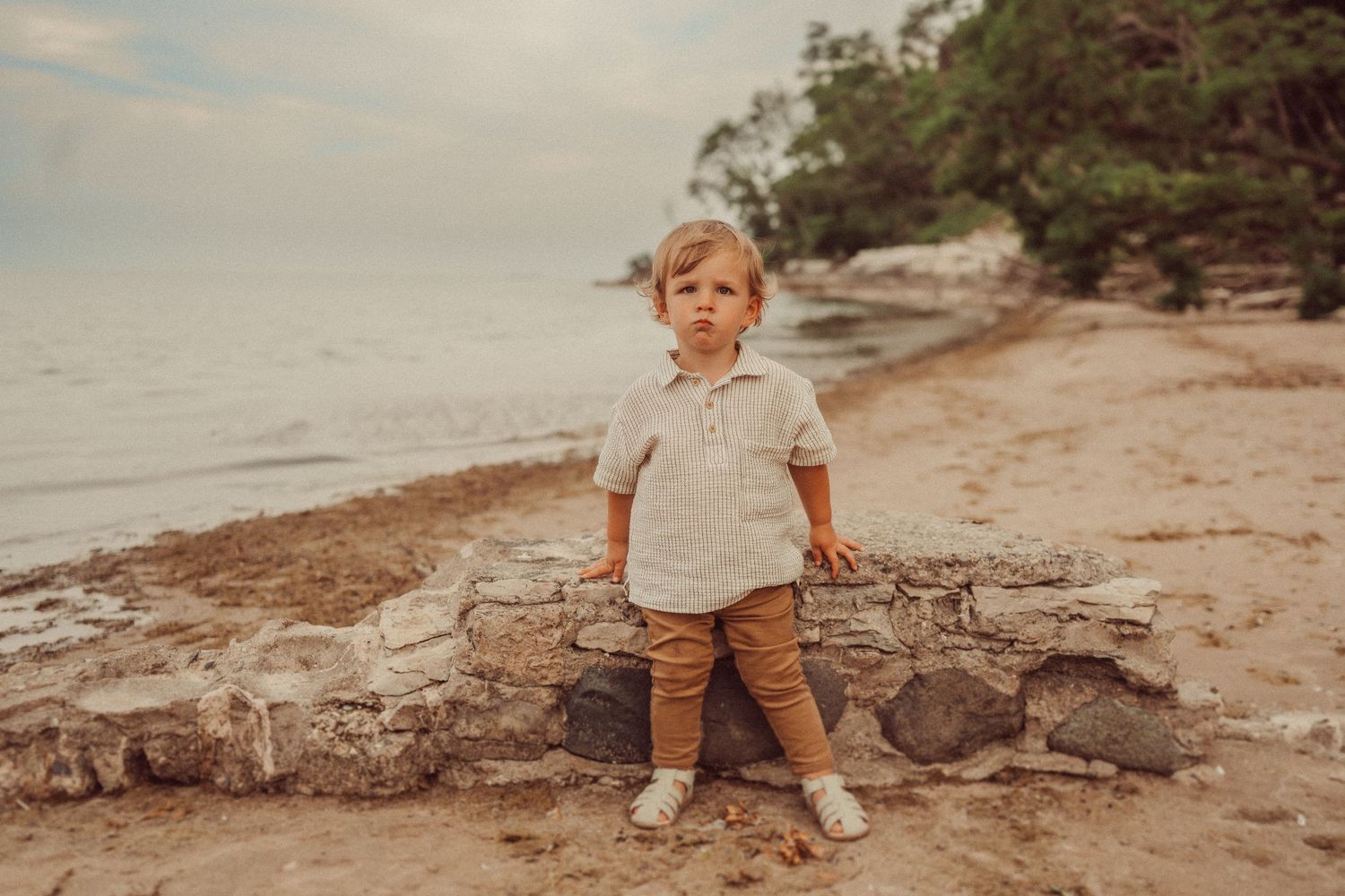 Small child in white shirt and brown pants stands on rocky beach shore.