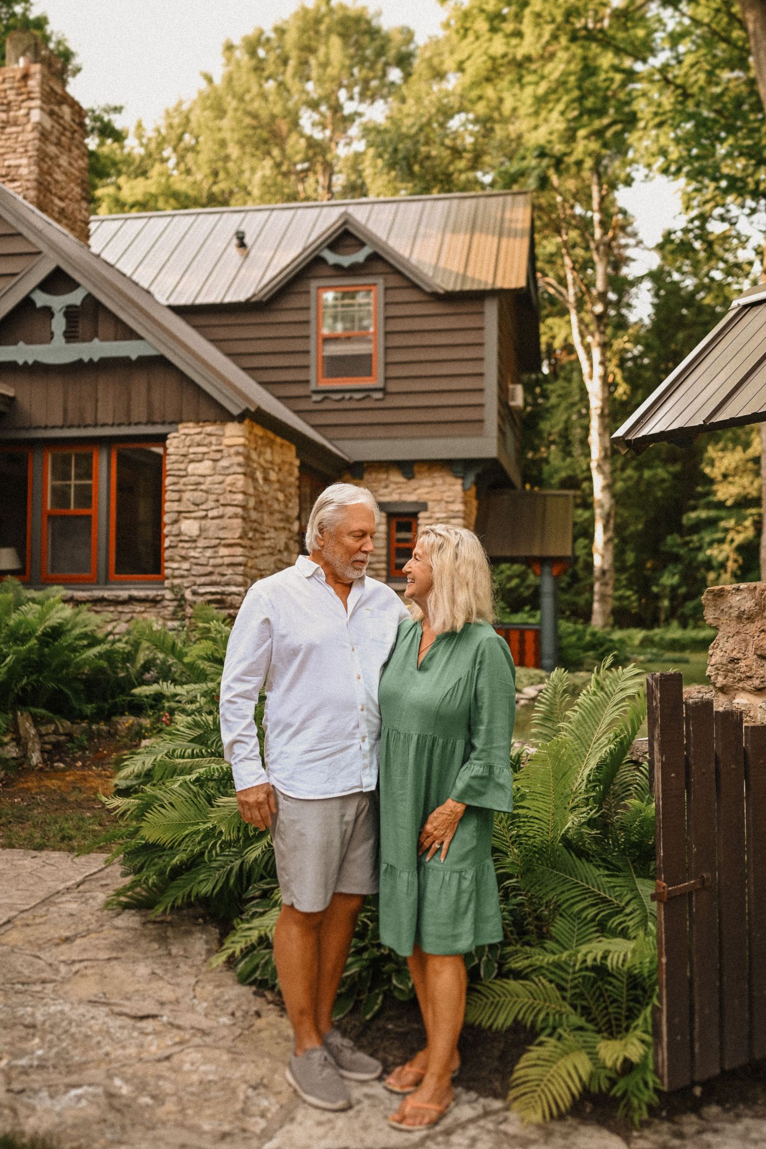 Couple in casual attire standing by stone pathway near ferns and wooden gate.