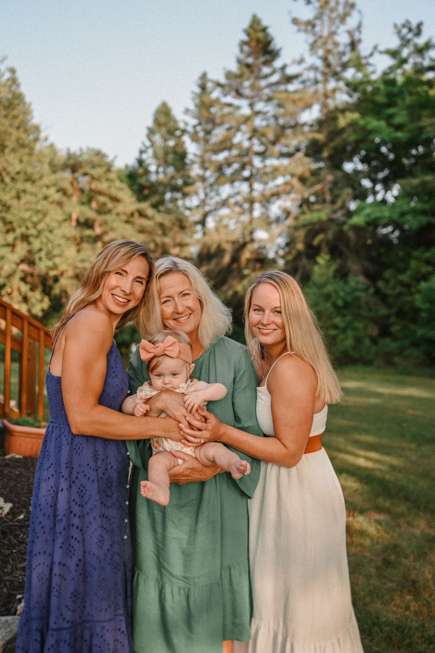 Three generations share loving smiles in a sunlit garden wearing summer dresses.