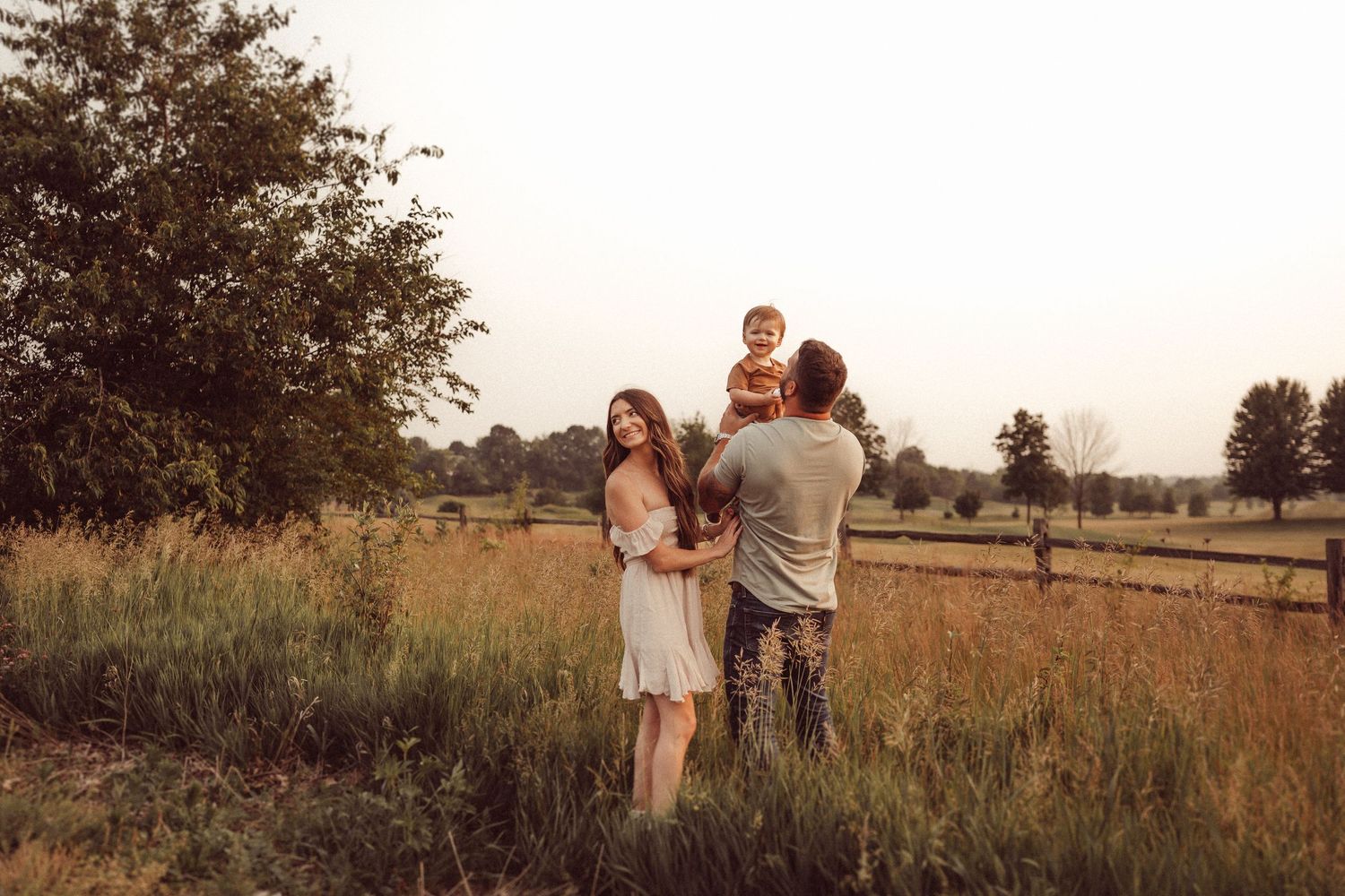 Family enjoying a warm sunset moment together in a rural field with wooden fencing in the background.