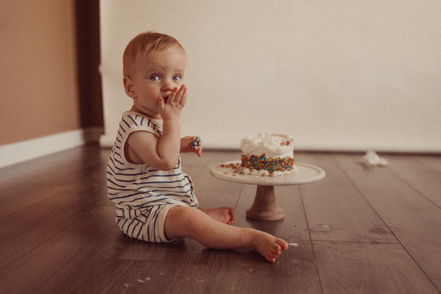 Side view of toddler in striped outfit sitting beside cake on wooden floor.