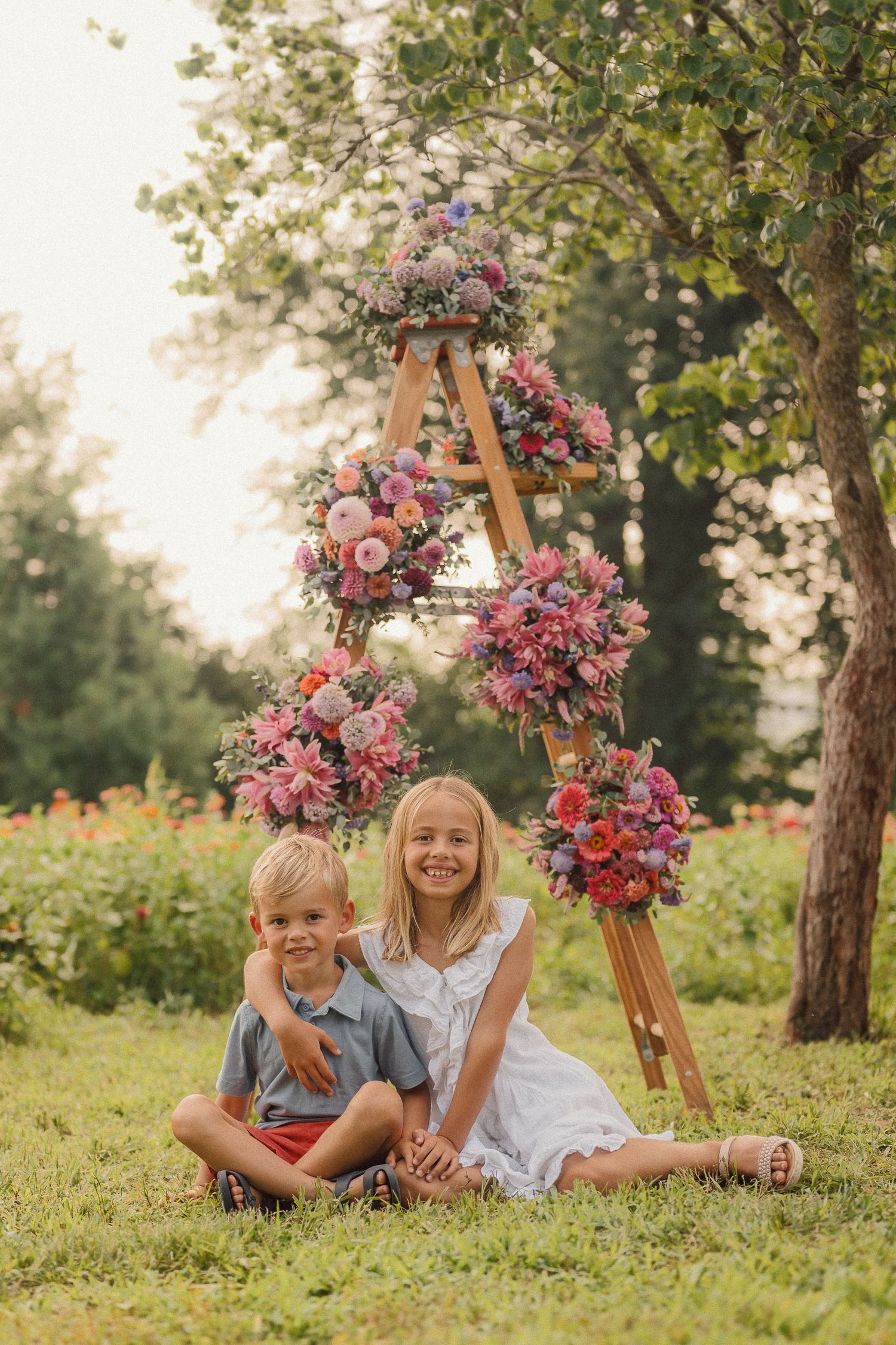 Rustic ladder adorned with pink flower bouquets stands against trees in a summer garden scene.
