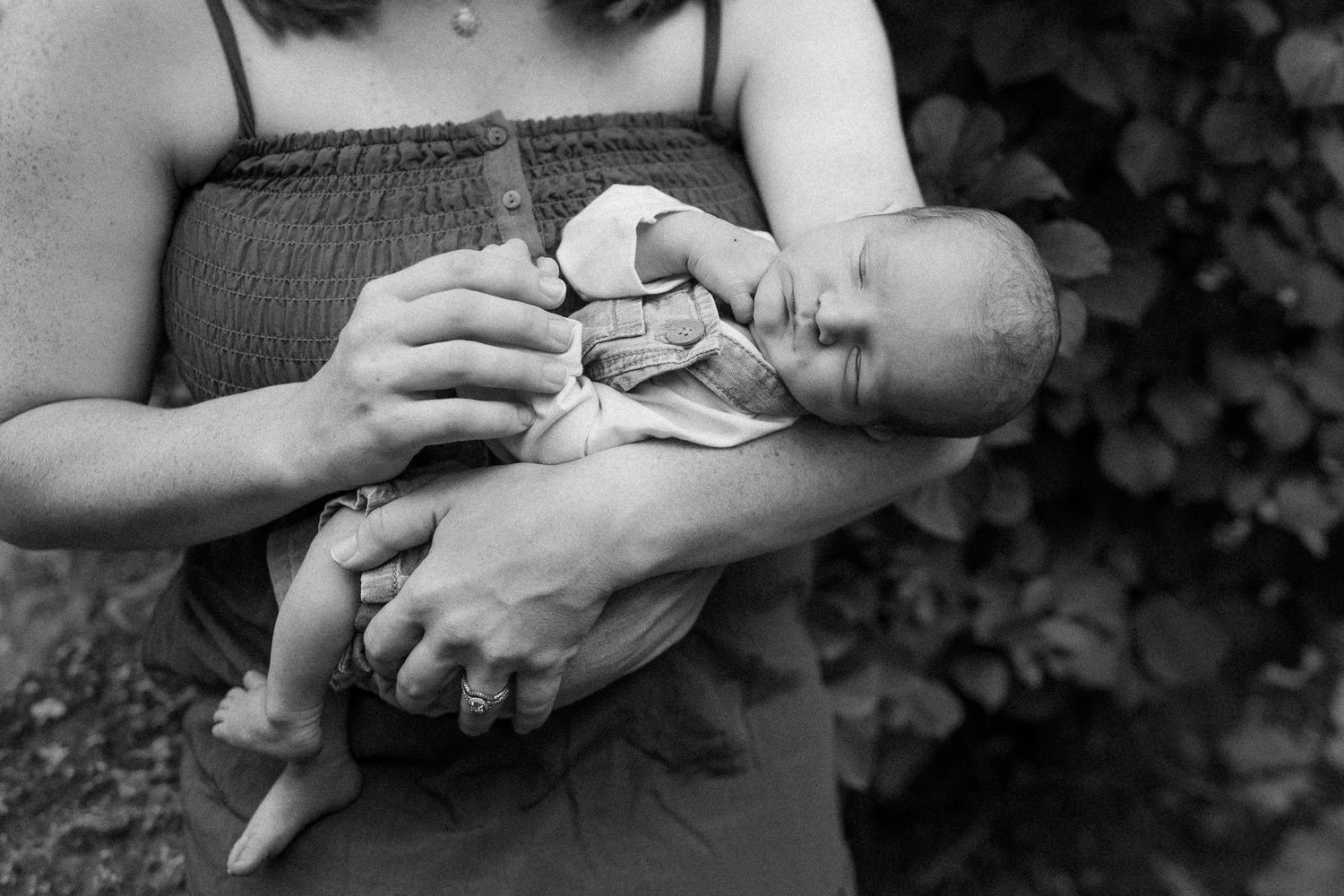 Black and white photo of hands holding a sleeping baby with soft shadows and leafy background.