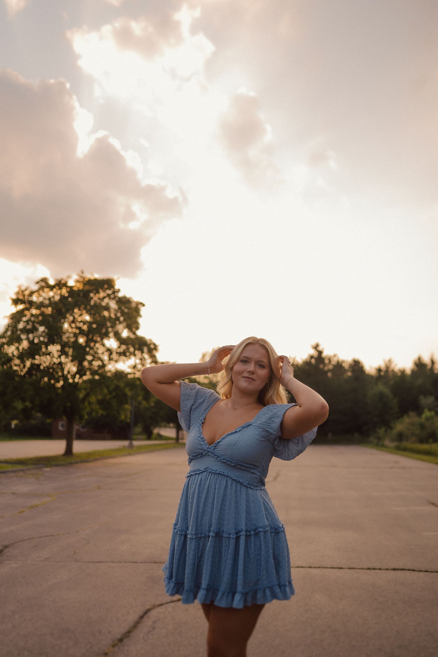 A figure in a blue dress poses expressively at sunset on an empty road surrounded by trees.