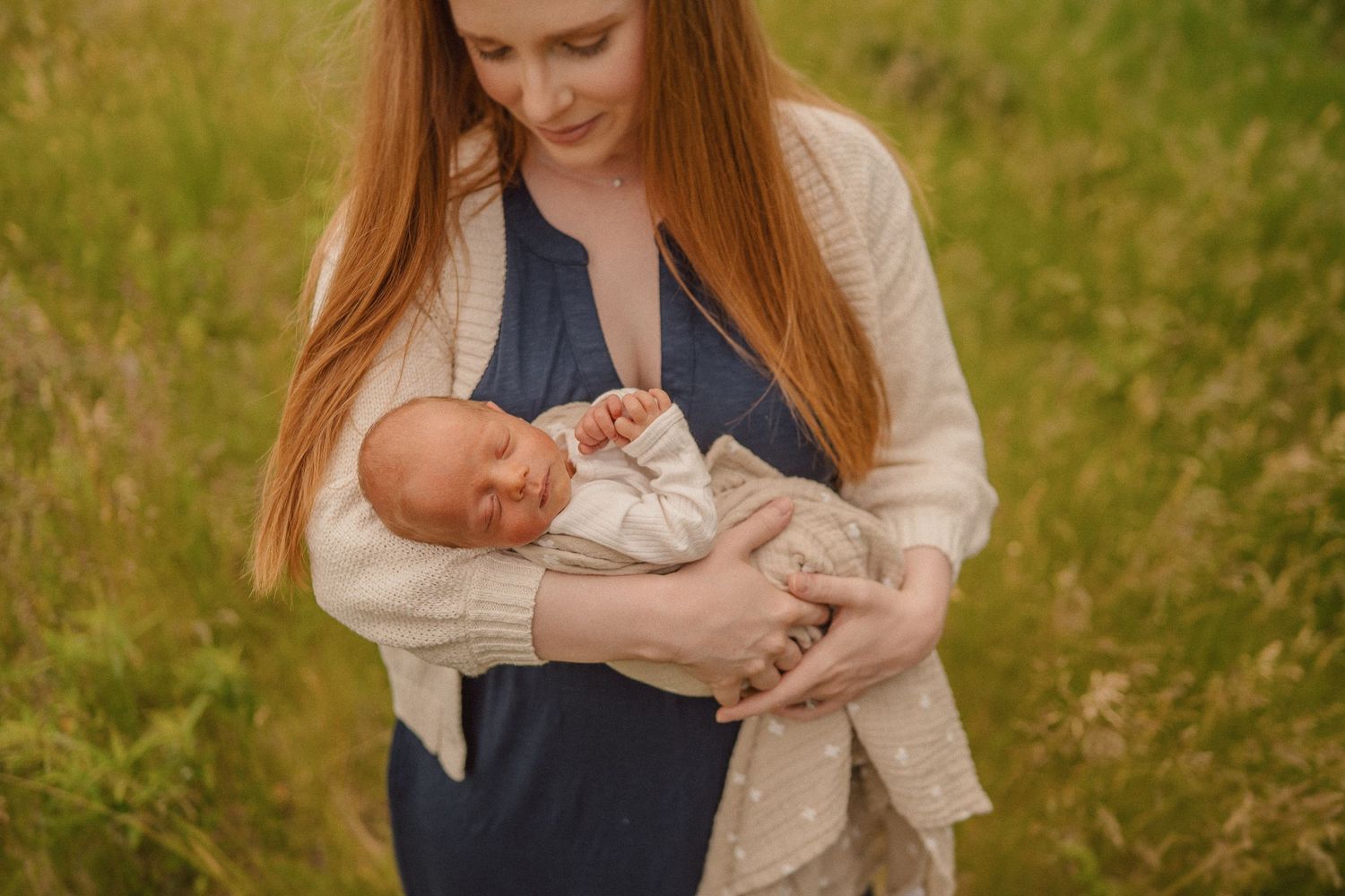 Woman in blue dress holds and cuddles baby outdoors in grassy field during golden hour.