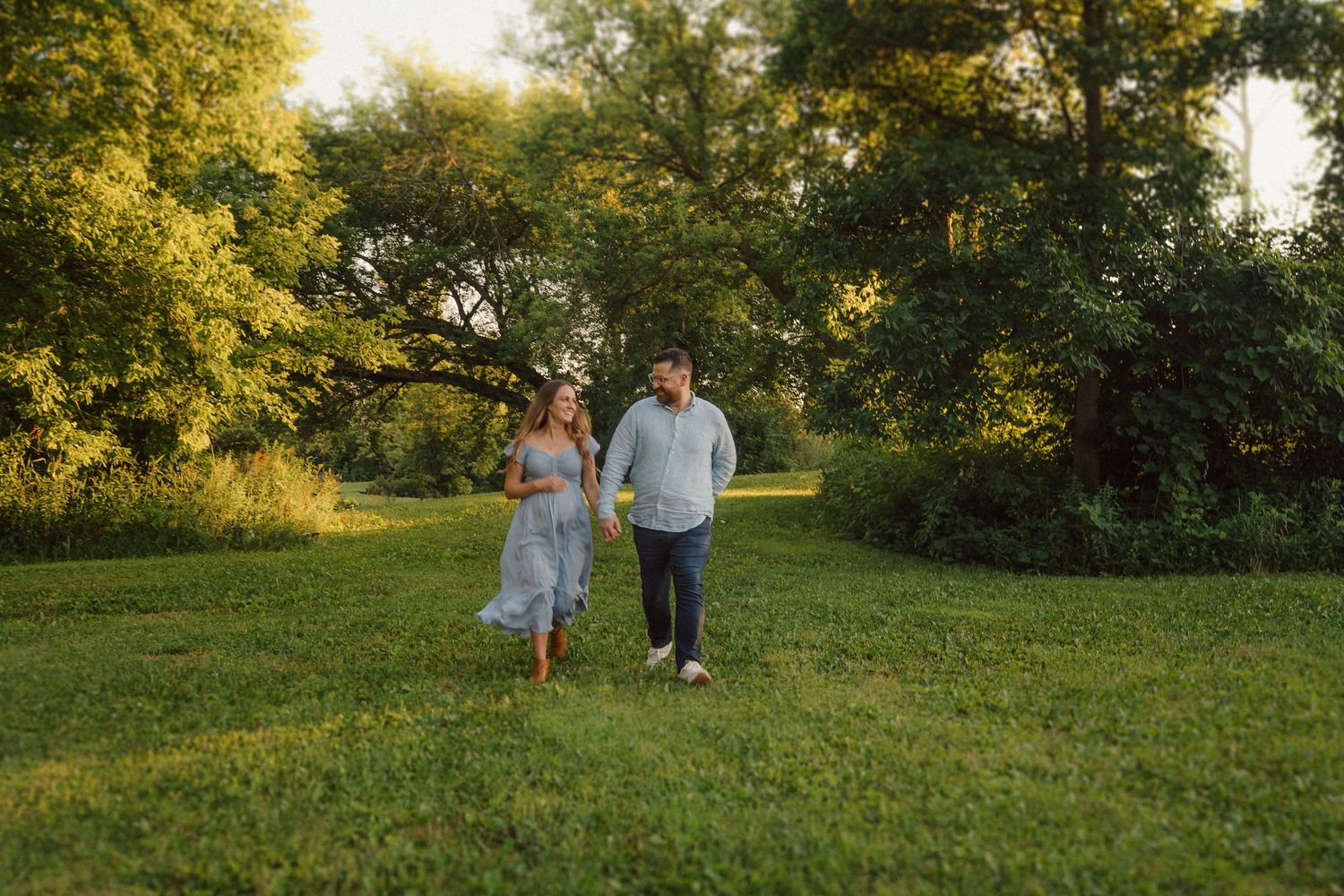 A couple walks hand in hand through a sunlit meadow during a romantic outdoor photo session.