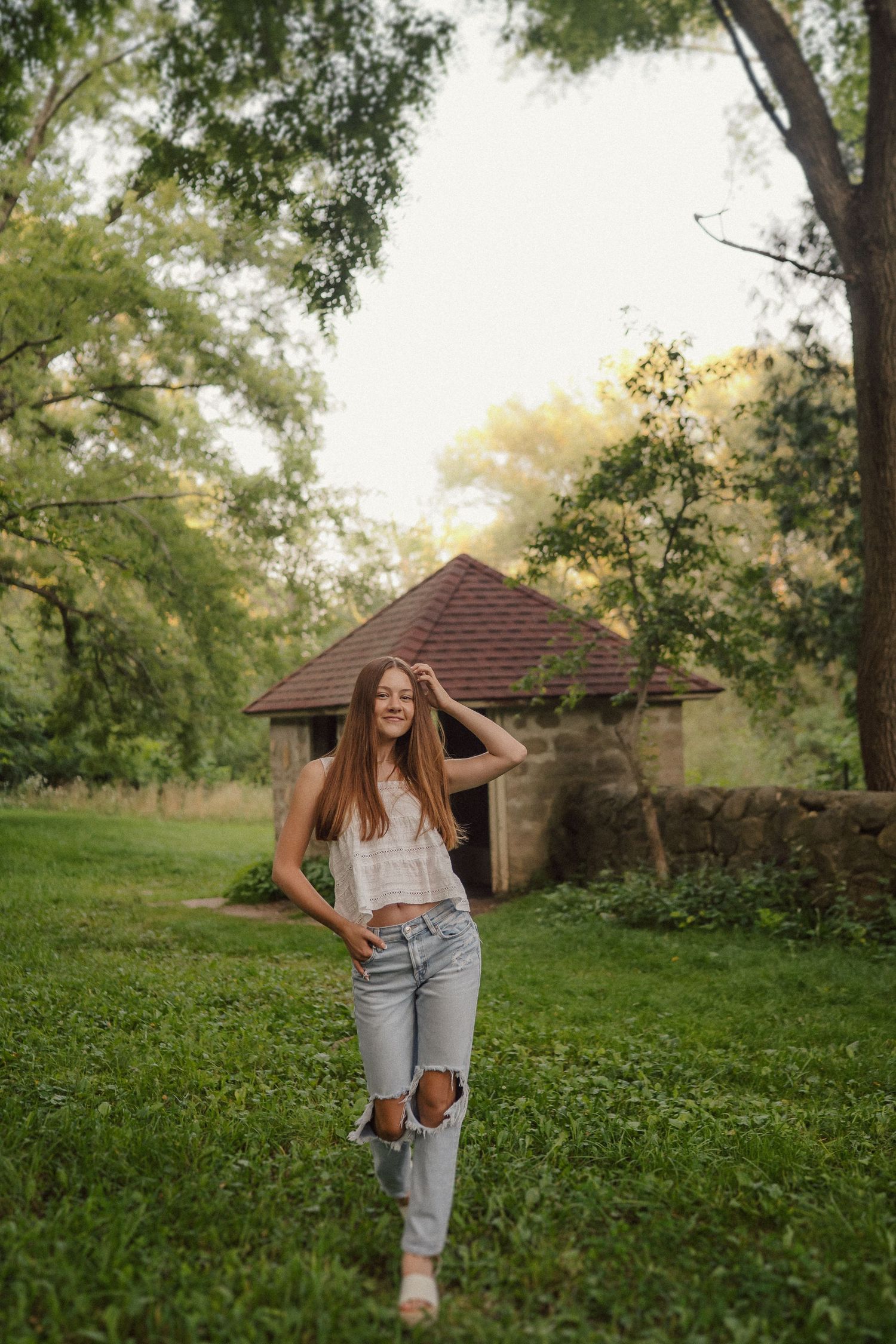 Person in distressed jeans and boots poses against lush greenery and rustic structures during golden hour.