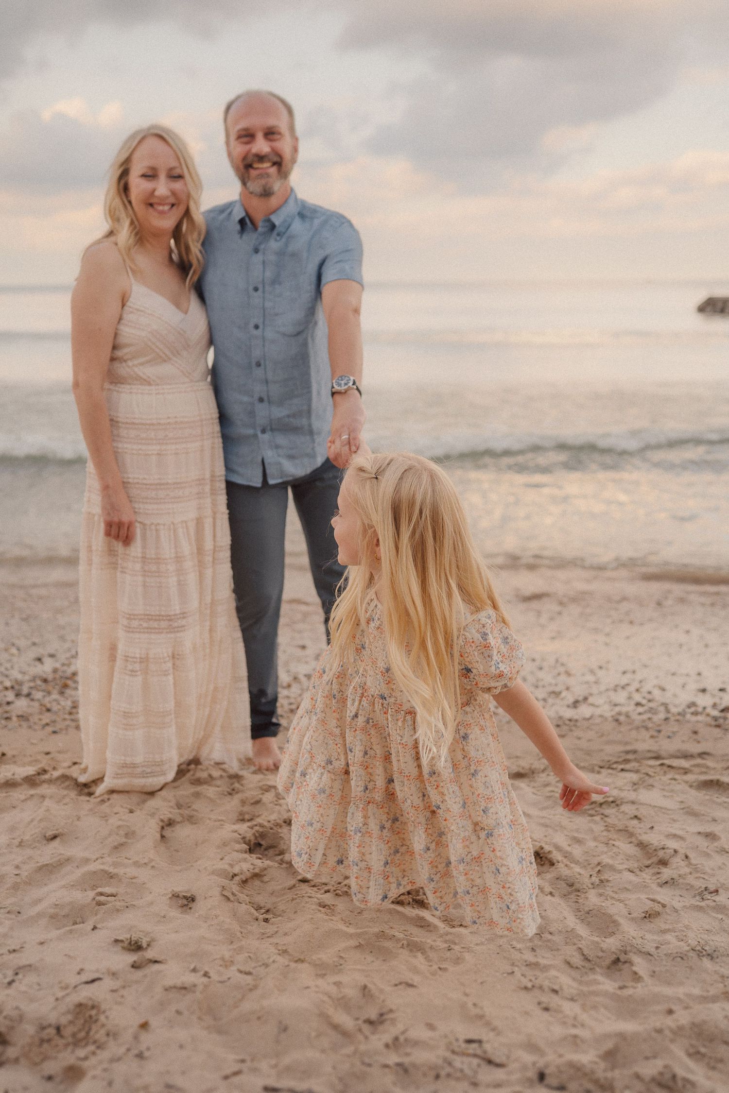 A family in coordinated cream and blue outfits poses together on a sandy beach at sunset.