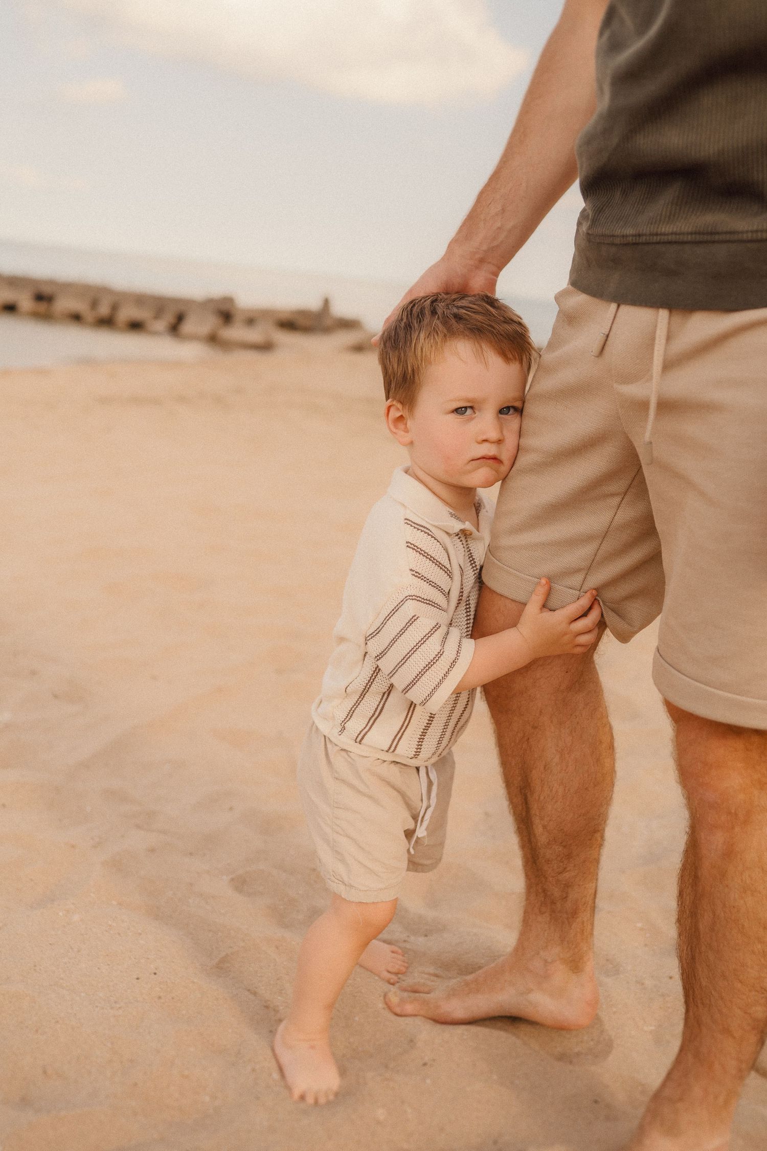 Small child stands beside adult feet on sandy beach while wearing cream colored clothing in warm natural lighting.