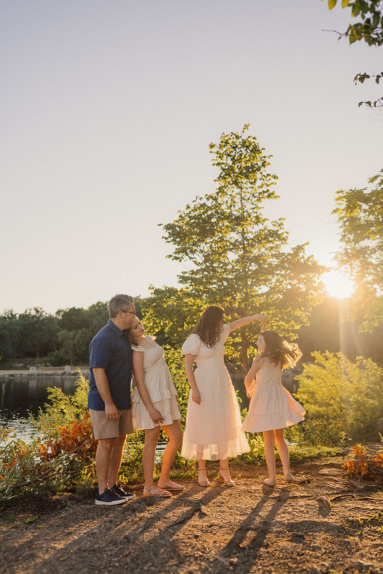 A family stands together on a hill during golden hour sunset with trees and a lake in the background.