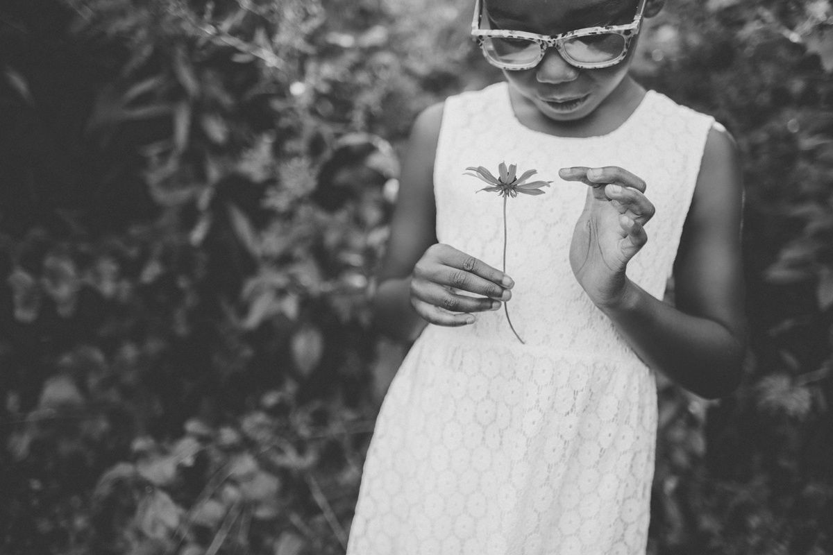 Black and white photo of a child in a white dress holding a flower in a garden setting.
