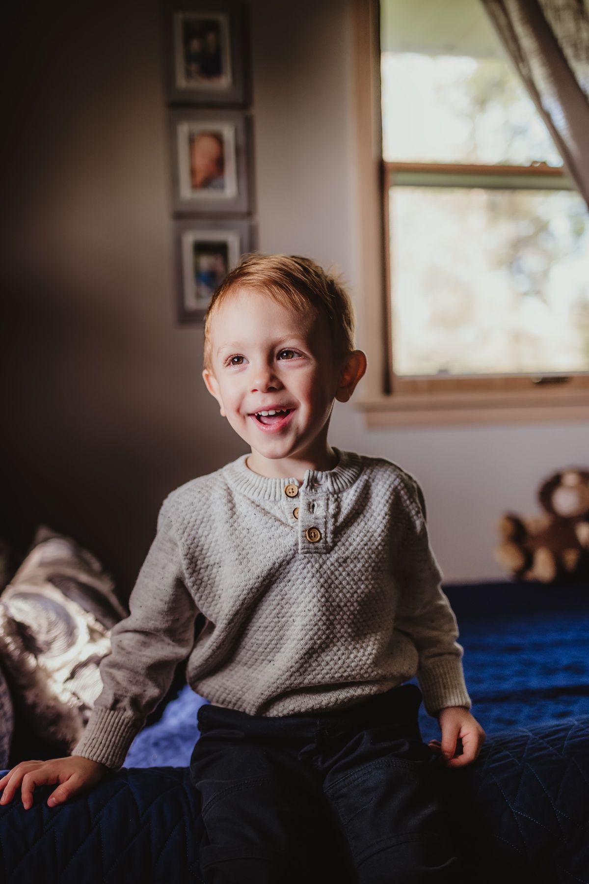 Child in gray knit sweater sits on blue bedding near window with framed photos on wall.