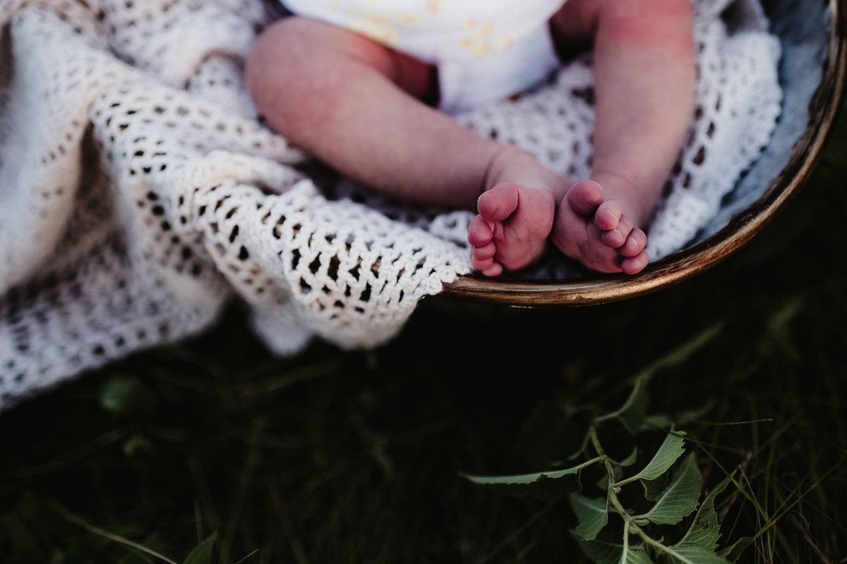 Tiny baby feet peek out from underneath a delicate white crocheted blanket against grass.