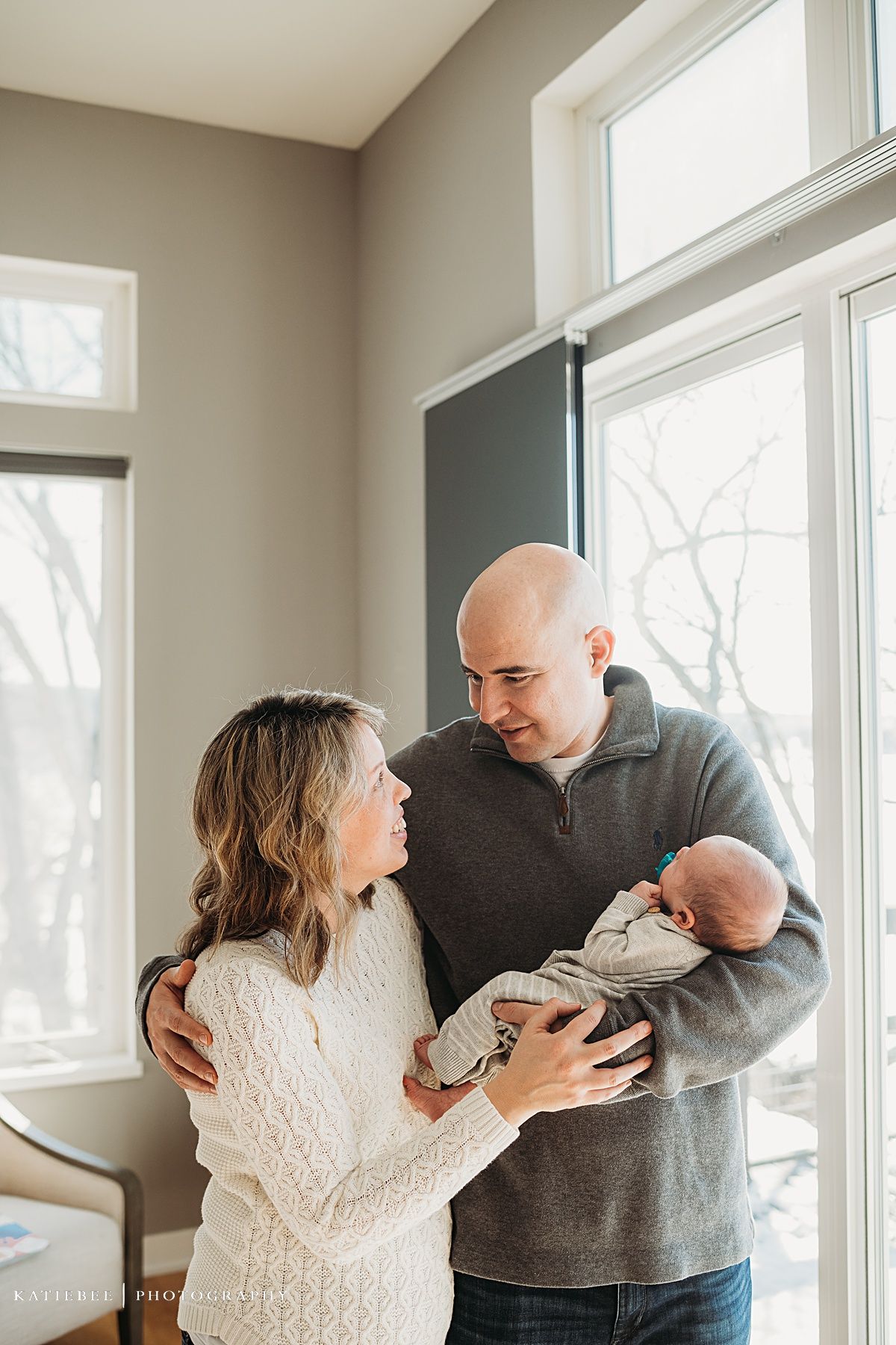 Intimate family moment by large windows as they gaze at their newborn baby.
